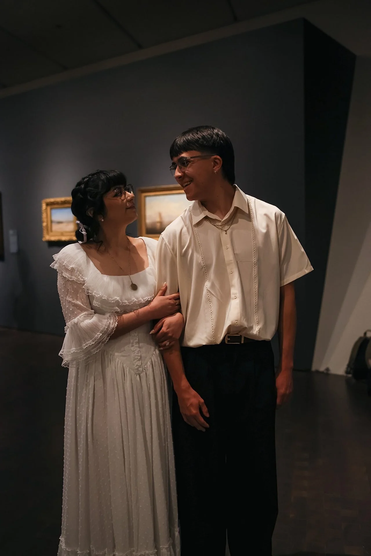 A candid portrait of a couple smiling at each other while walking through a museum hallway, surrounded by framed paintings for relaxed city engagement photos with movement.