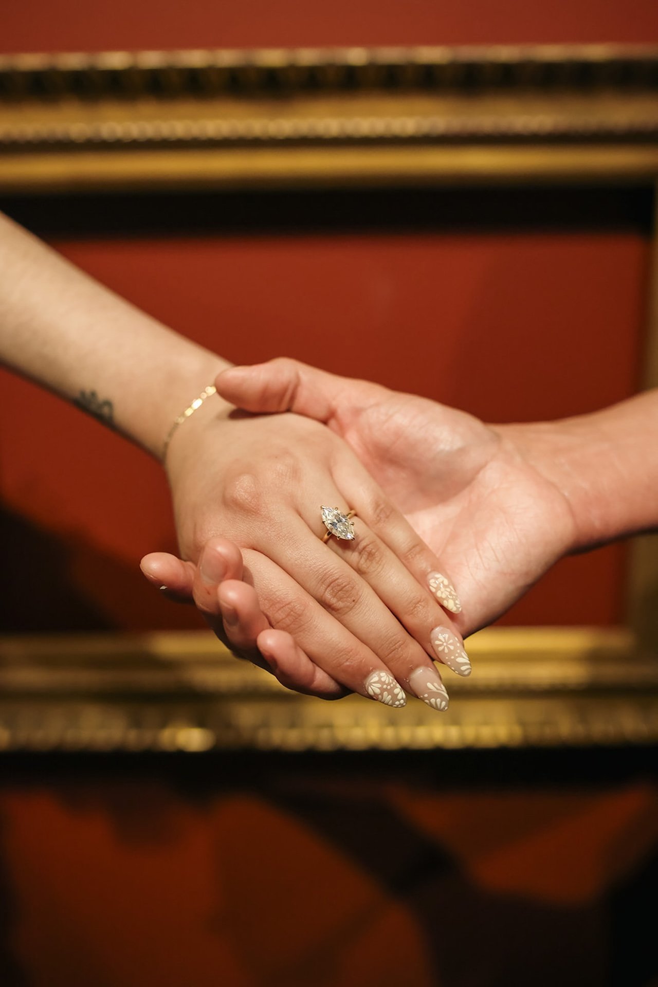 A close-up of two hands intertwined, highlighting an engagement ring with soft museum lighting and warm-toned frames blurred in the background.