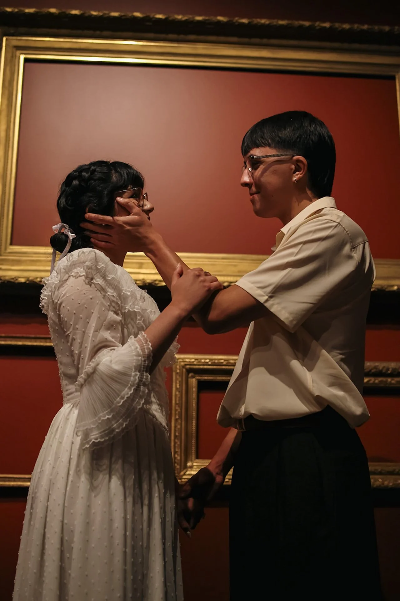 A tender moment of a couple standing face to face in front of deep red gallery walls and ornate gold frames, hands gently holding each other, creating intimate city engagement photos inside a museum.