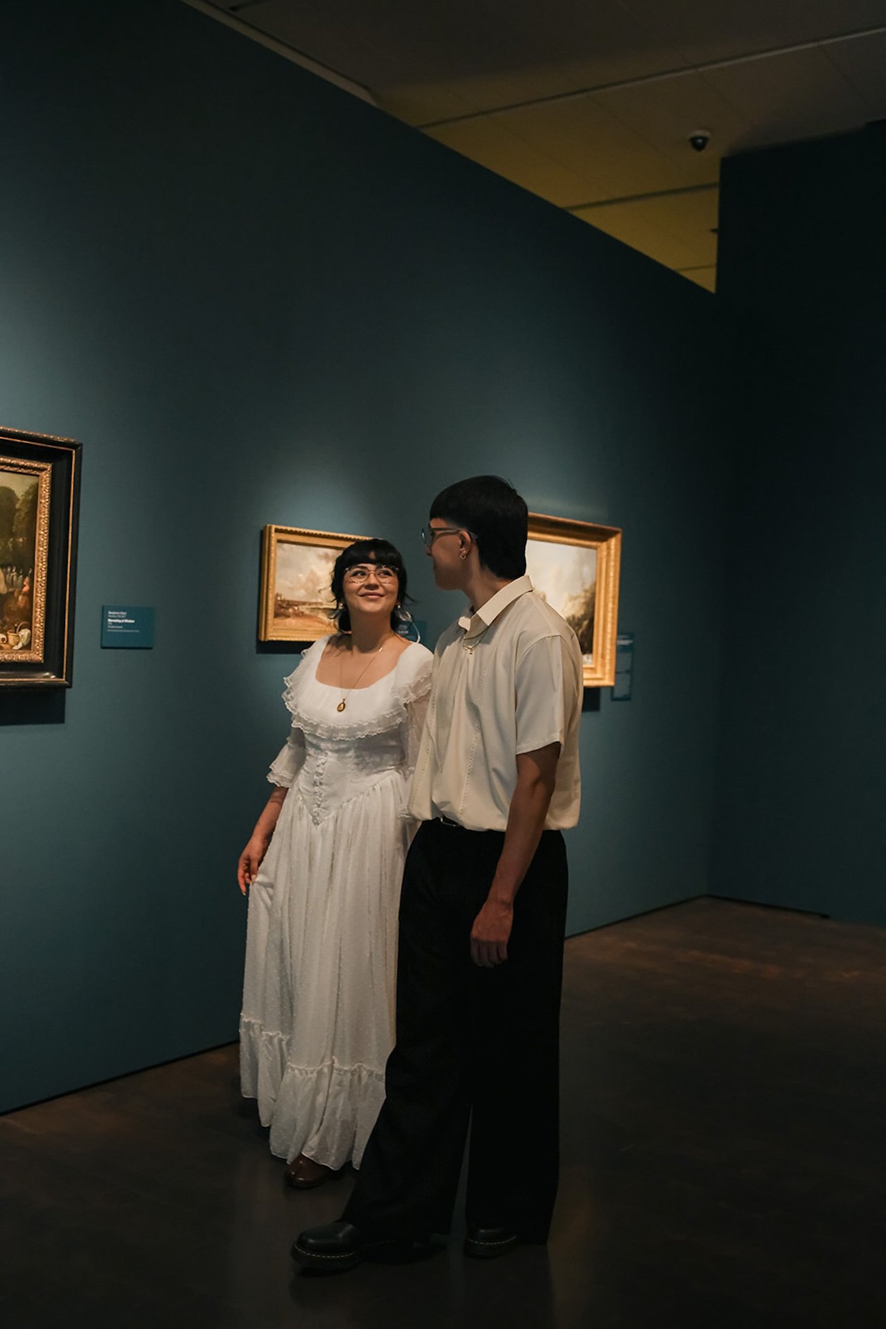 A candid walking moment of a couple strolling hand in hand through a museum hallway with teal walls and framed paintings, showcasing relaxed city engagement photos.