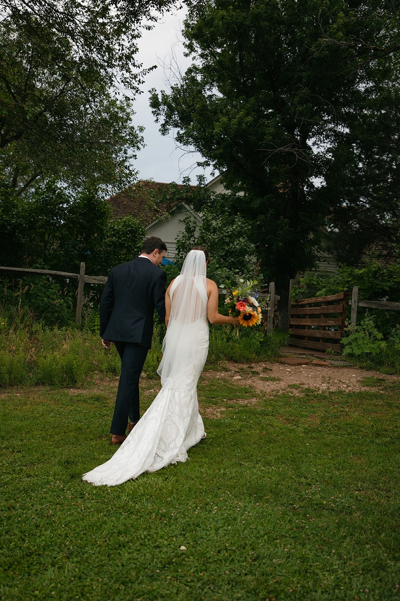 Bride and groom walking away hand in hand with the bride’s long lace train flowing behind her in a rustic outdoor setting.