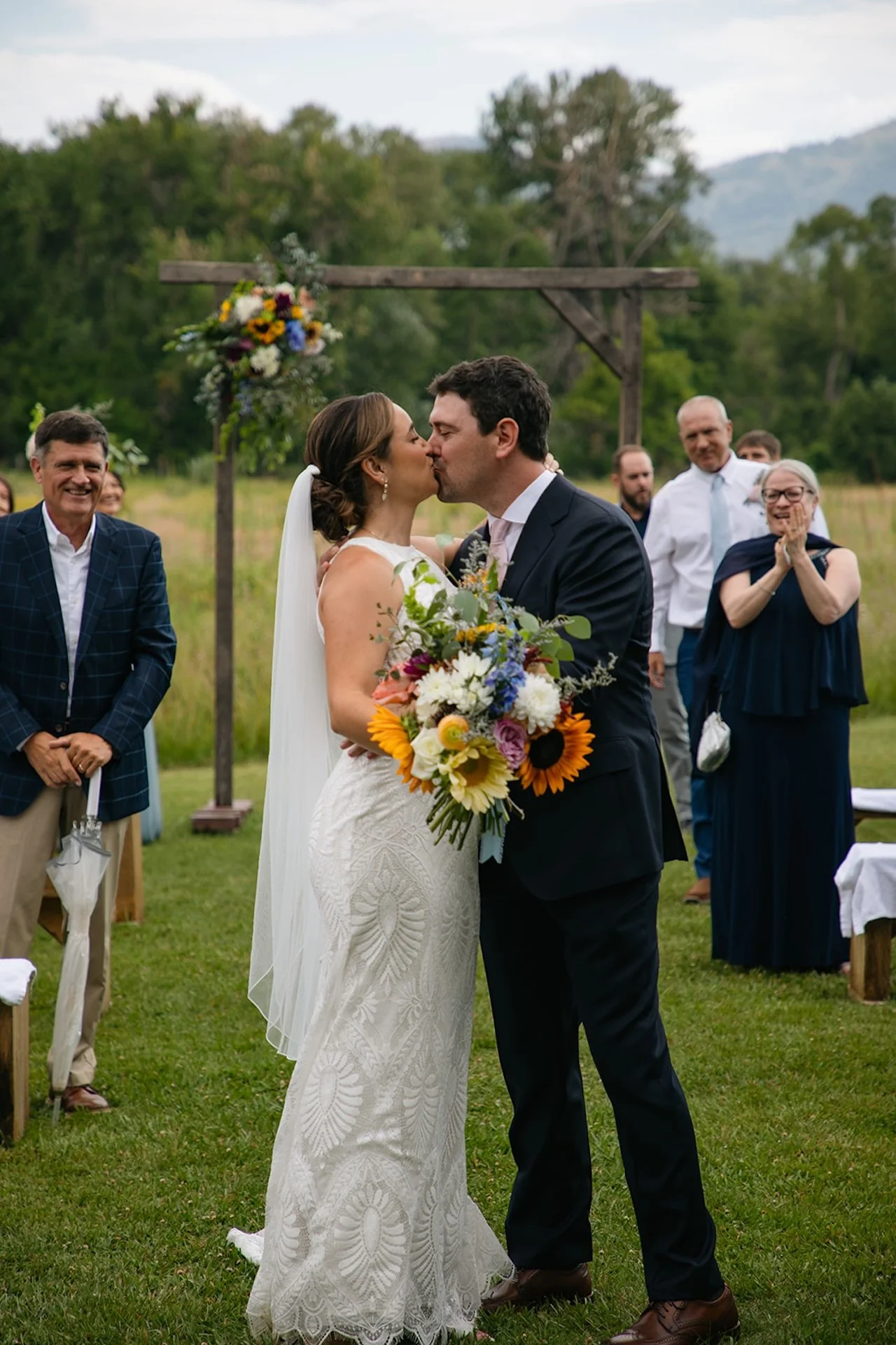 Bride and groom sharing their first kiss at the ceremony while guests clap and smile in the background.