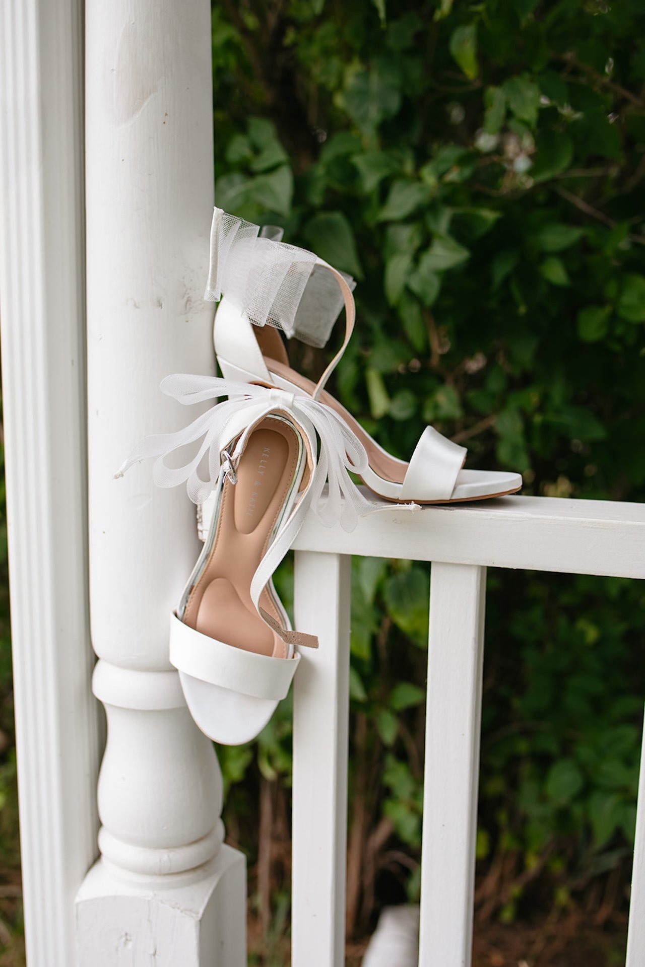 White bridal heels with delicate tulle bows styled on a porch railing for wedding detail photos.