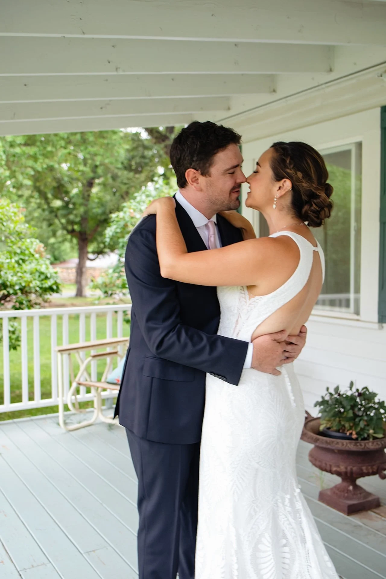Bride and groom embracing and nearly kissing on a light blue porch with greenery and white railing in the background.