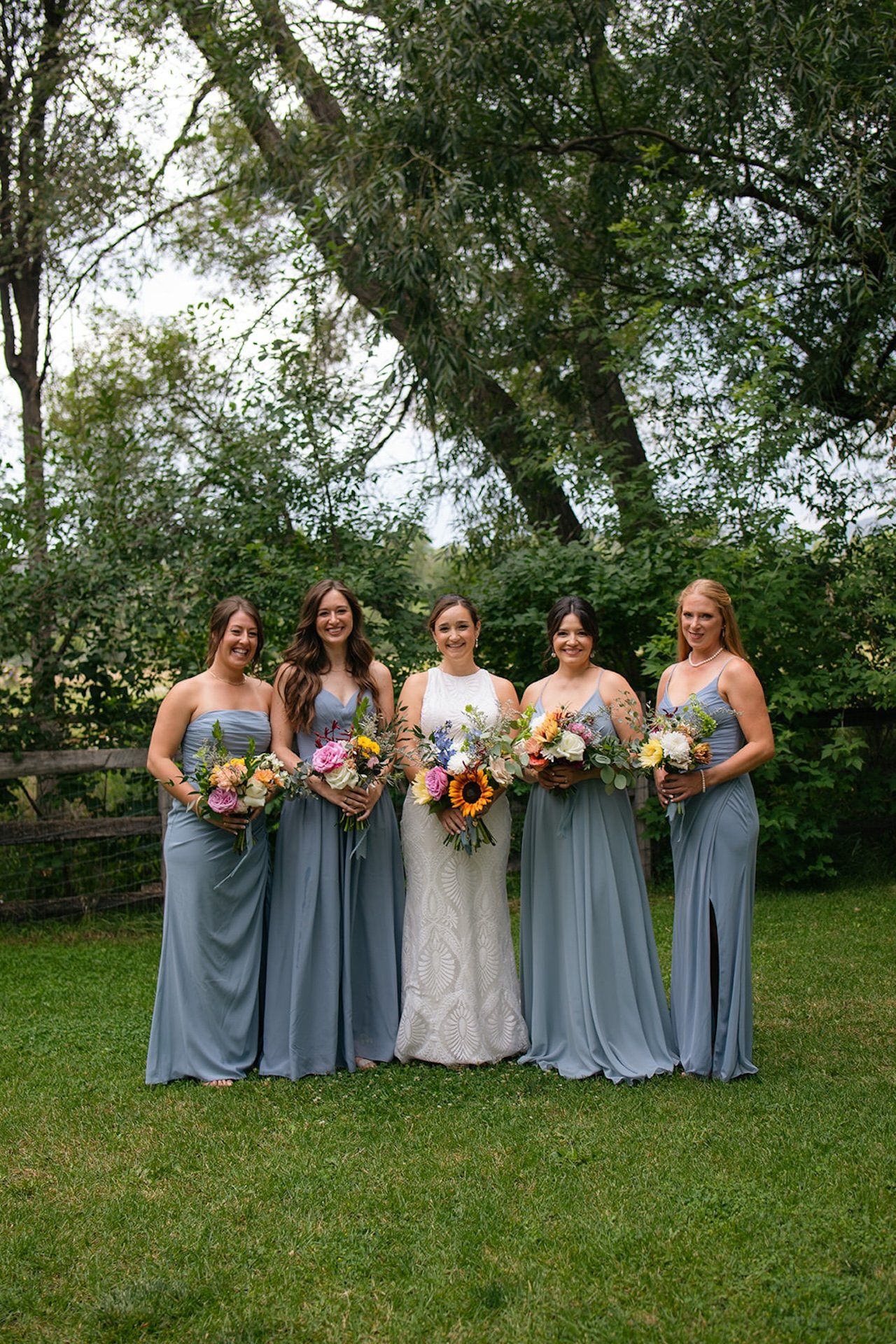 Bride standing with bridesmaids in soft blue dresses holding colorful bouquets at a Fort Collins Wedding Venue with lush trees and a rustic fence backdrop.
