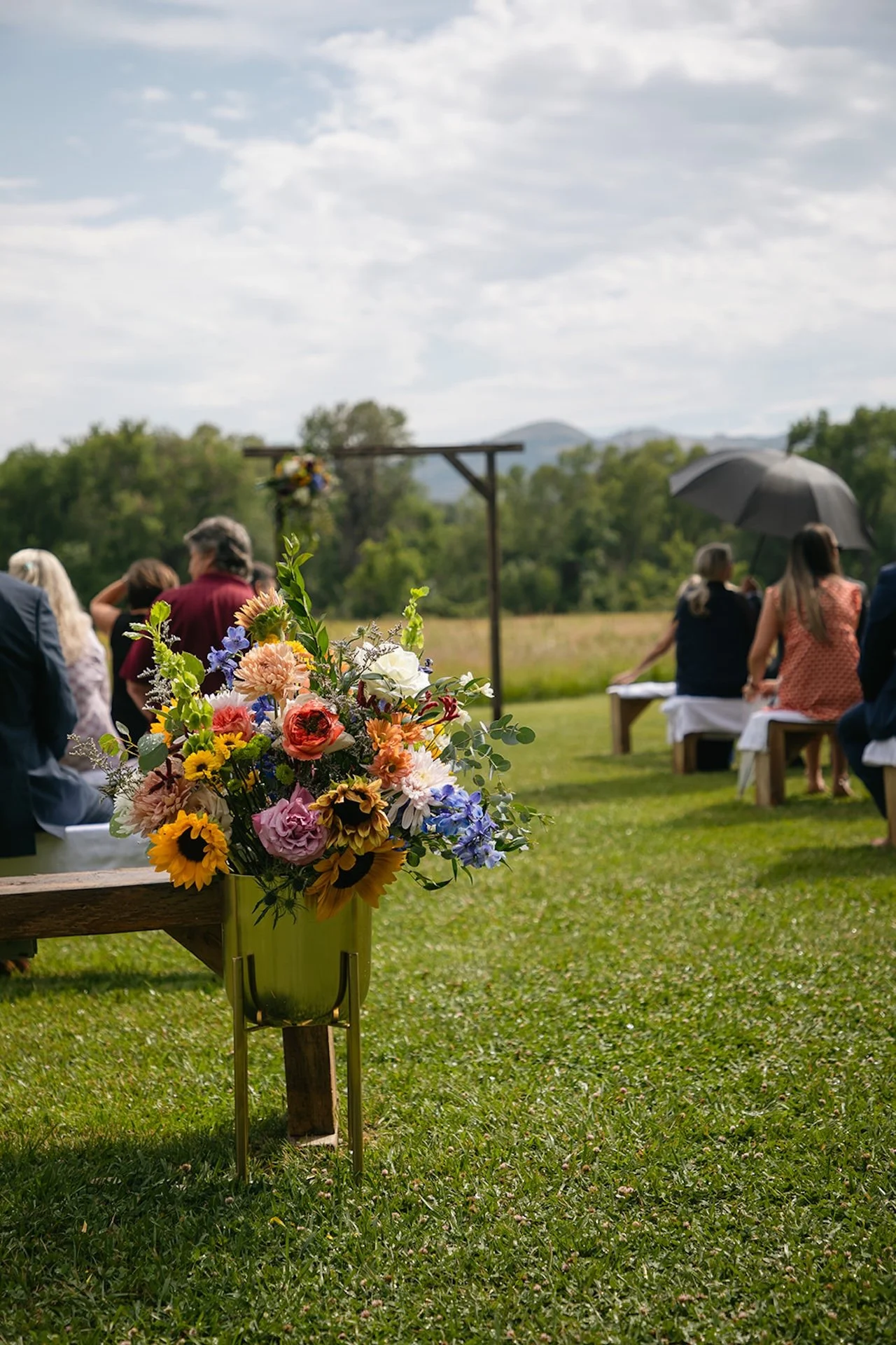 Outdoor wedding ceremony setup with wooden benches, floral arrangements, and a wooden arbor in a grassy field with mountain views.