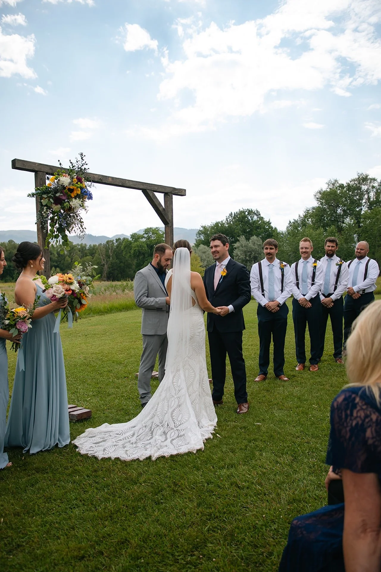 Bride and groom exchanging vows under a wooden ceremony arbor with colorful florals during an outdoor Fort Collins Wedding Venue ceremony surrounded by greenery and mountains.