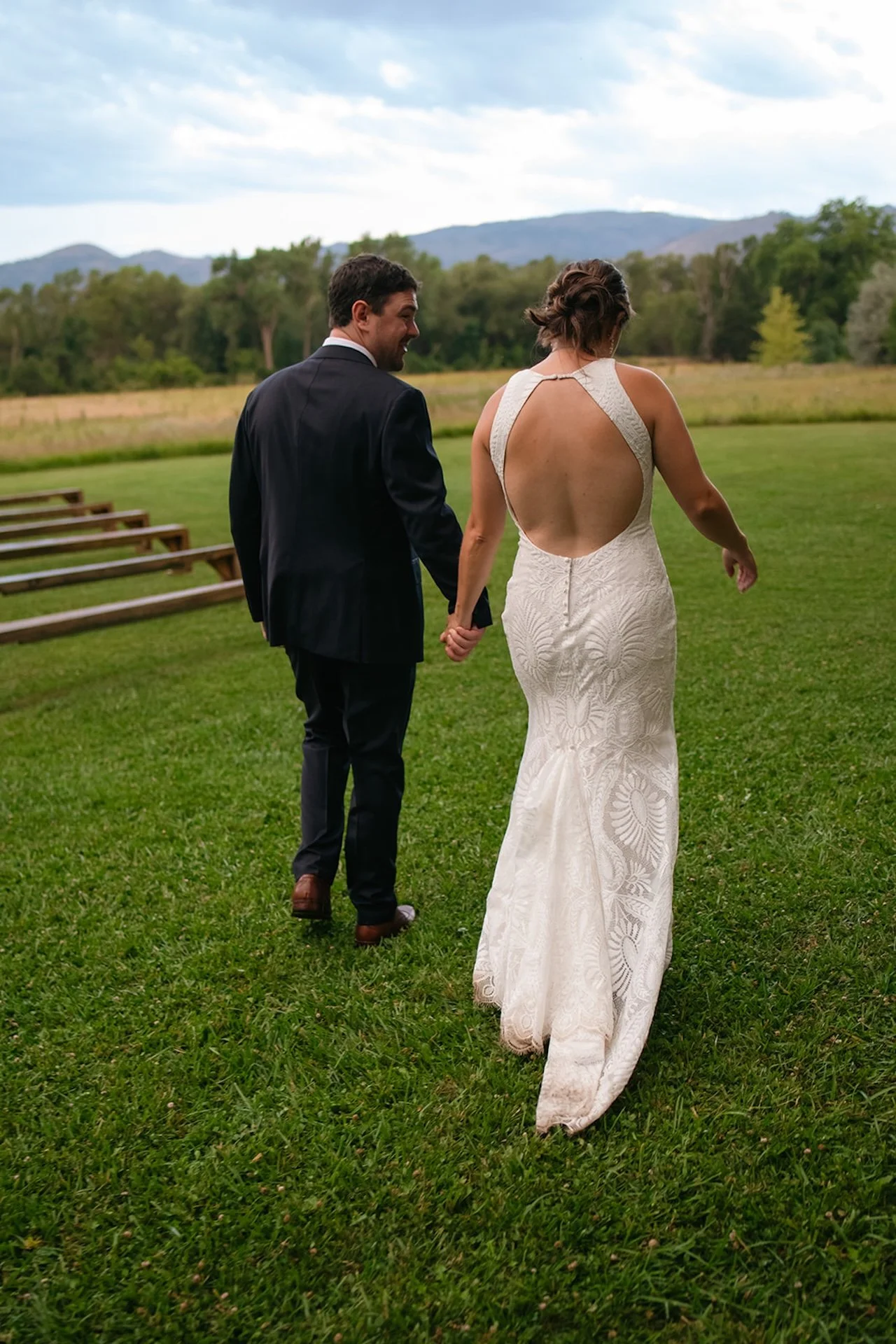 Bride and groom walking hand in hand across a grassy field with mountains and trees in the background, showing the open-back wedding dress.
