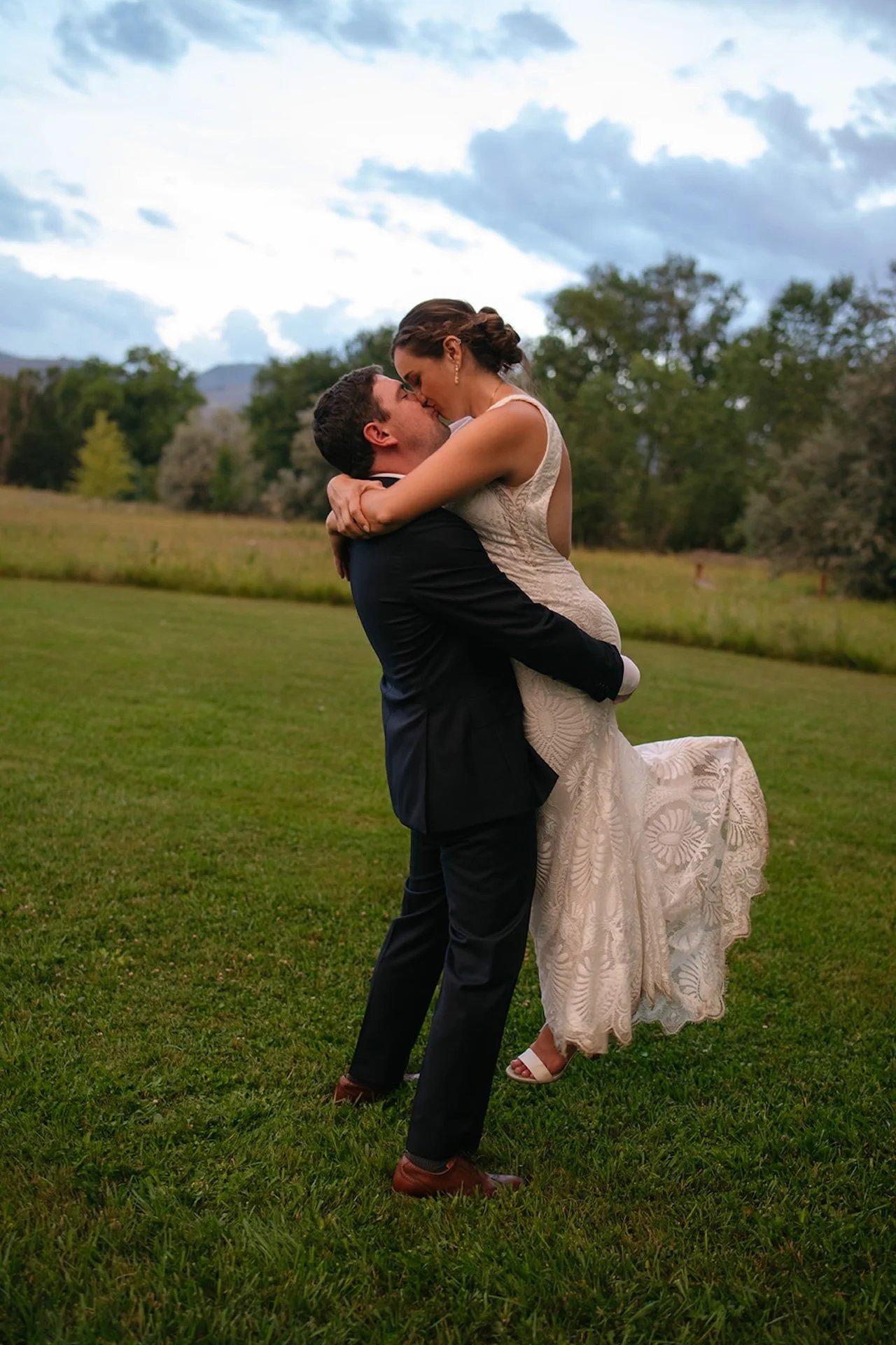 Groom lifting the bride in a field while they share a kiss during golden hour with mountains and trees in the distance.