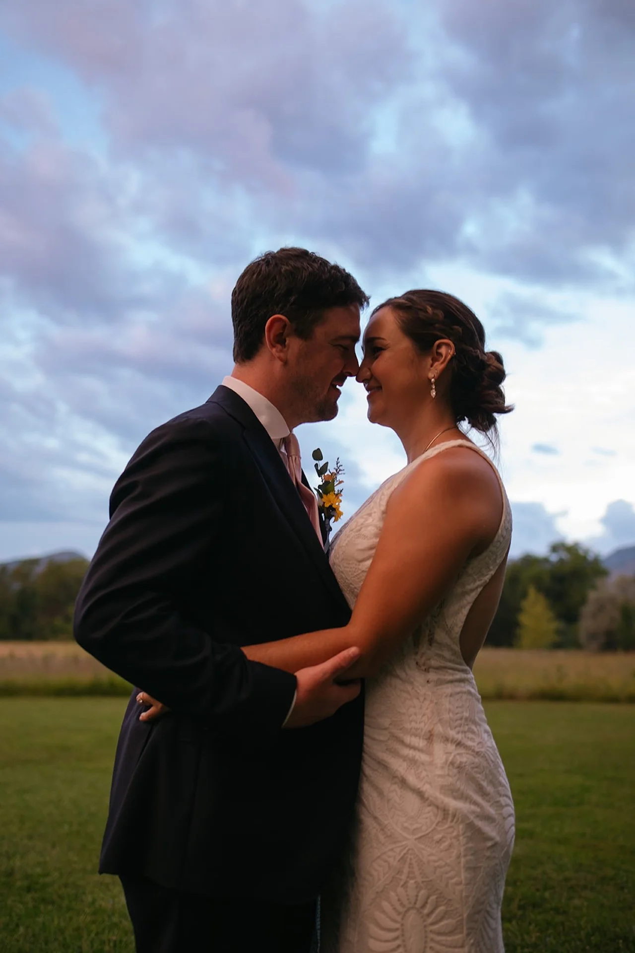 Bride and groom standing nose to nose in a field at sunset, embracing with mountains and a dramatic cloudy sky in the background.