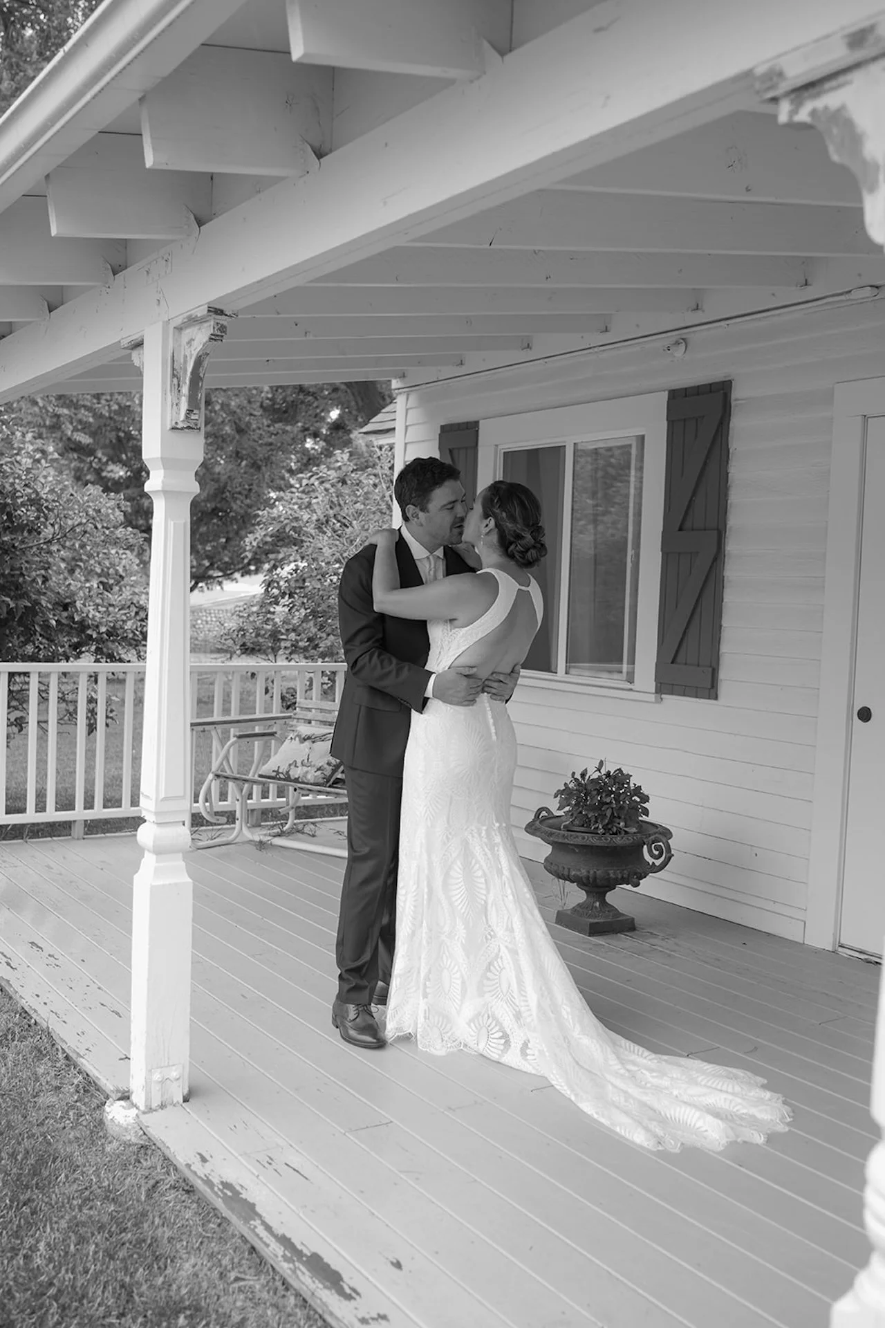 Bride and groom embracing on a white farmhouse porch with wooden beams, railings, and greenery in a black and white portrait.