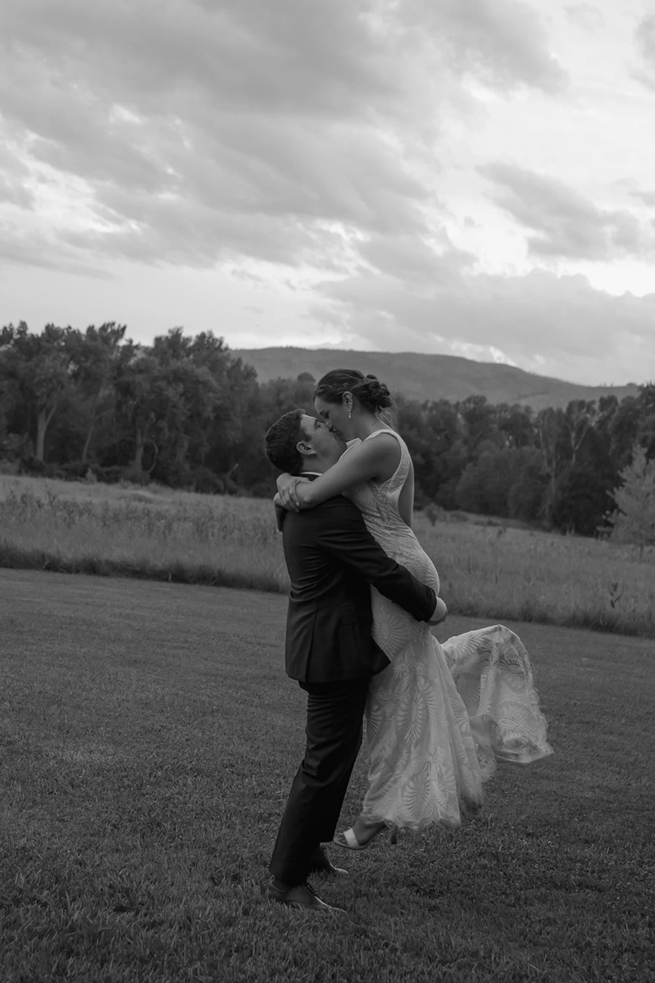 Groom lifting the bride in a field while they kiss, with her lace dress flowing and mountains in the background in a black and white portrait.