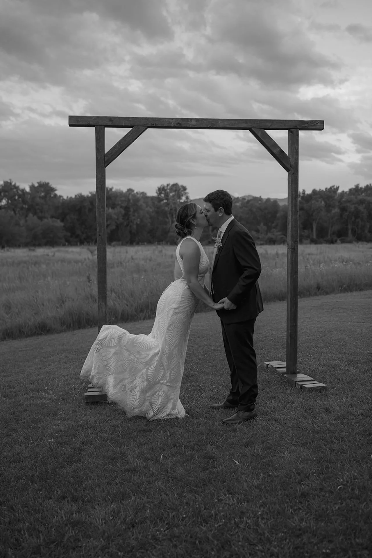 Bride and groom holding hands and leaning in for a kiss under a wooden arbor with dramatic cloudy skies in a black and white portrait.
