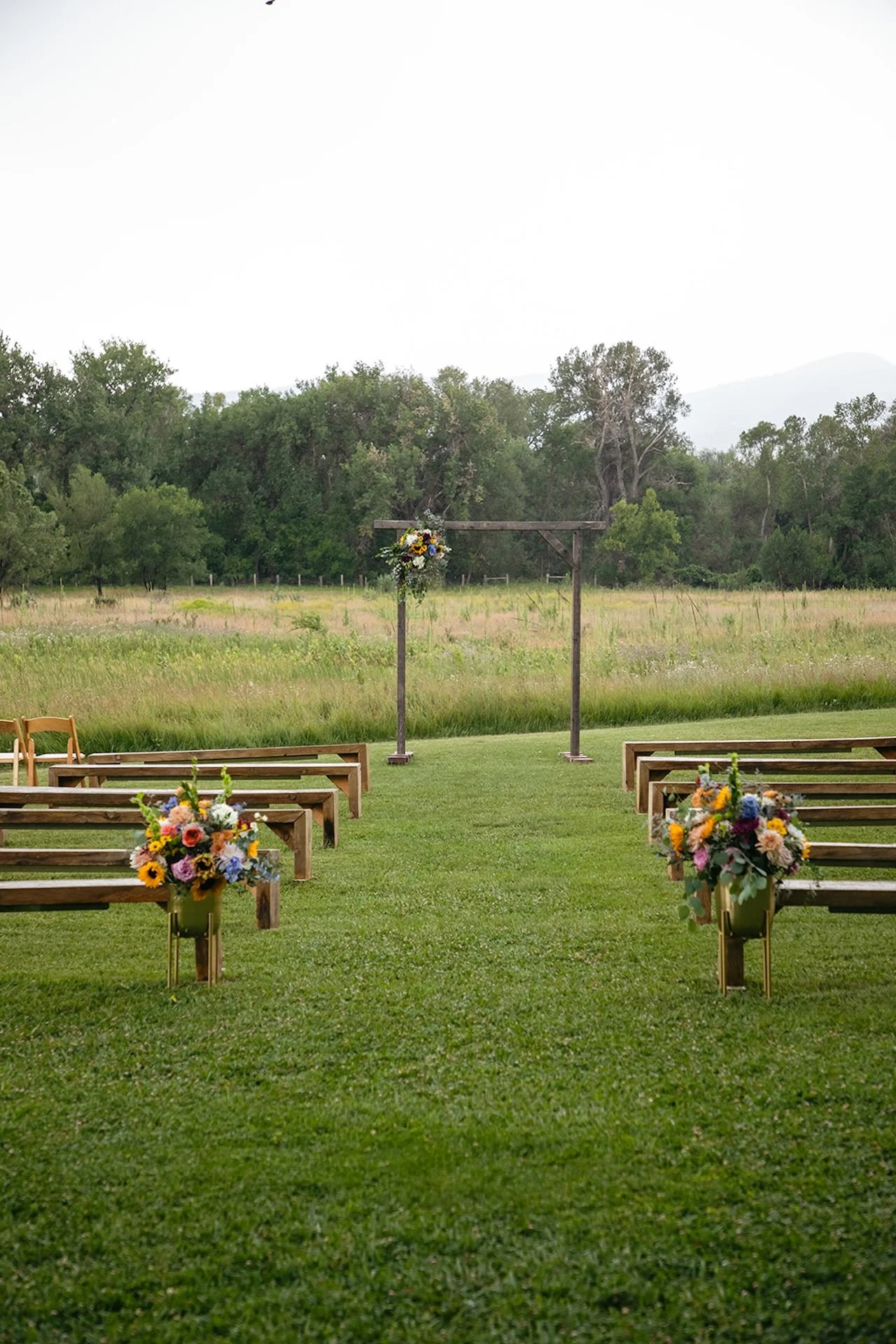 Empty outdoor wedding ceremony setup with wooden benches, floral arrangements in gold stands, and a wooden arbor in a grassy field.