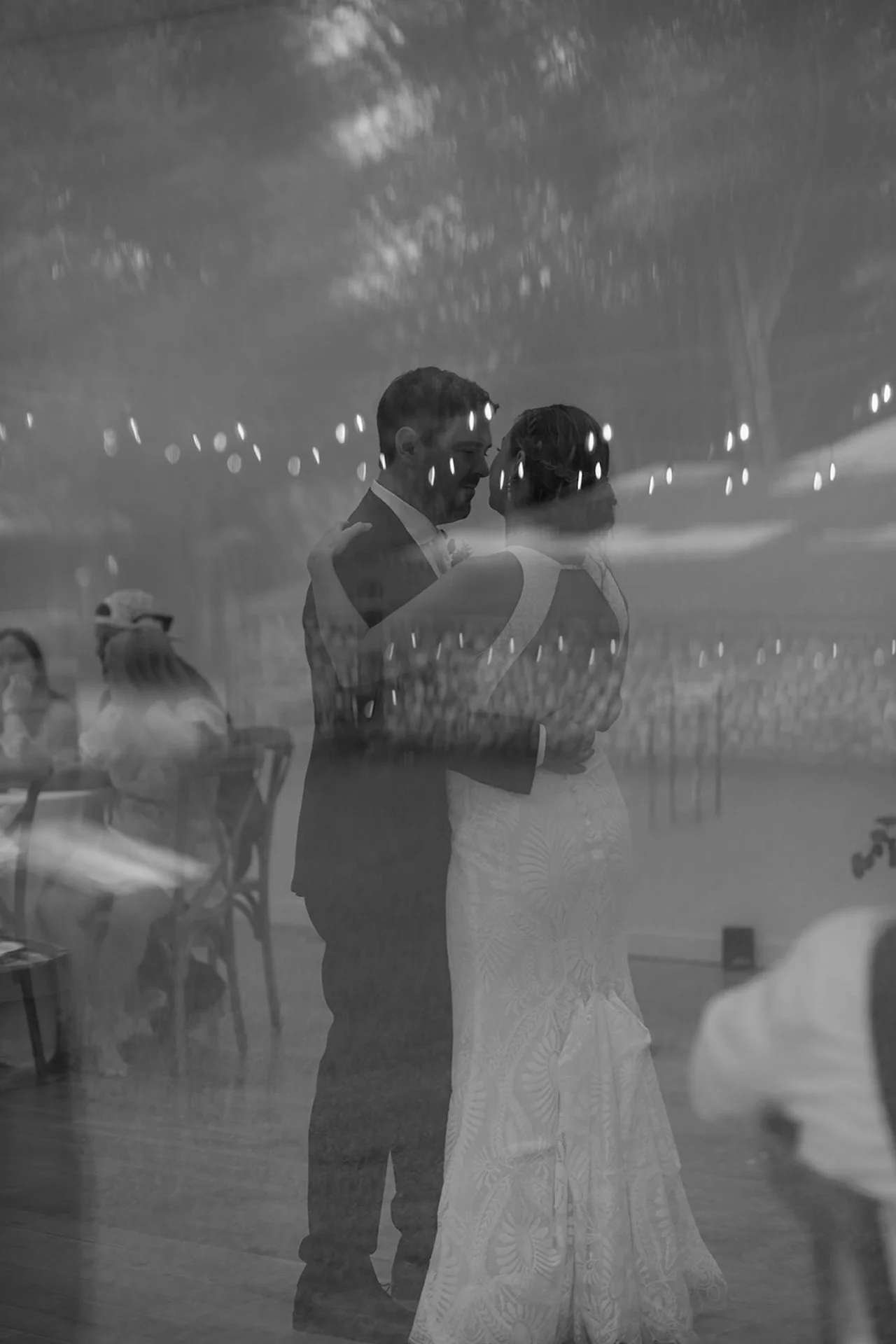 Bride and groom dancing closely during their first dance with reflections and string lights visible through a glass window for a dreamy layered effect.