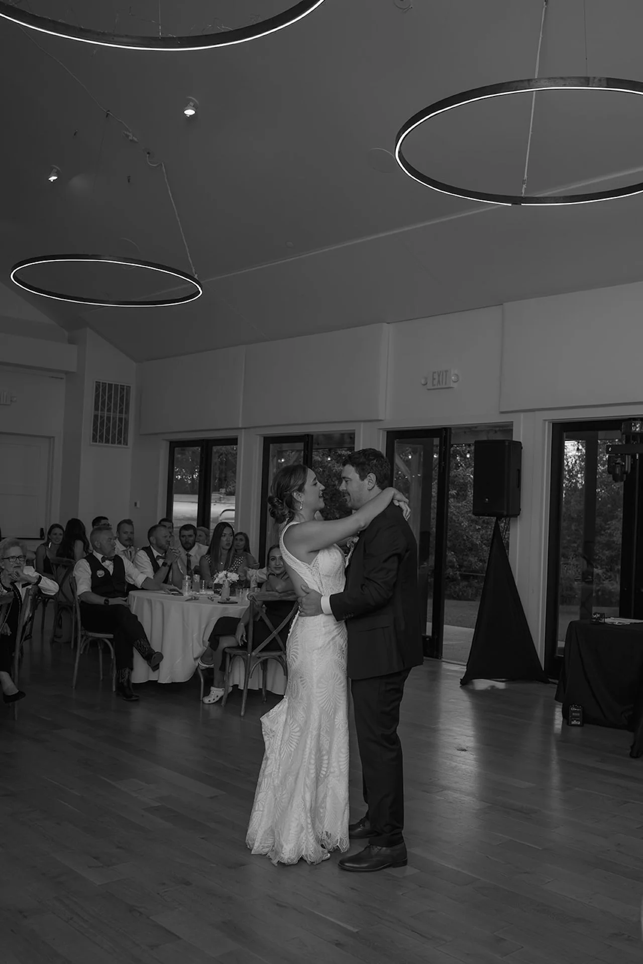 Bride and groom slow dancing indoors during their wedding reception with modern circular ceiling lights and guests seated at round tables in the background.