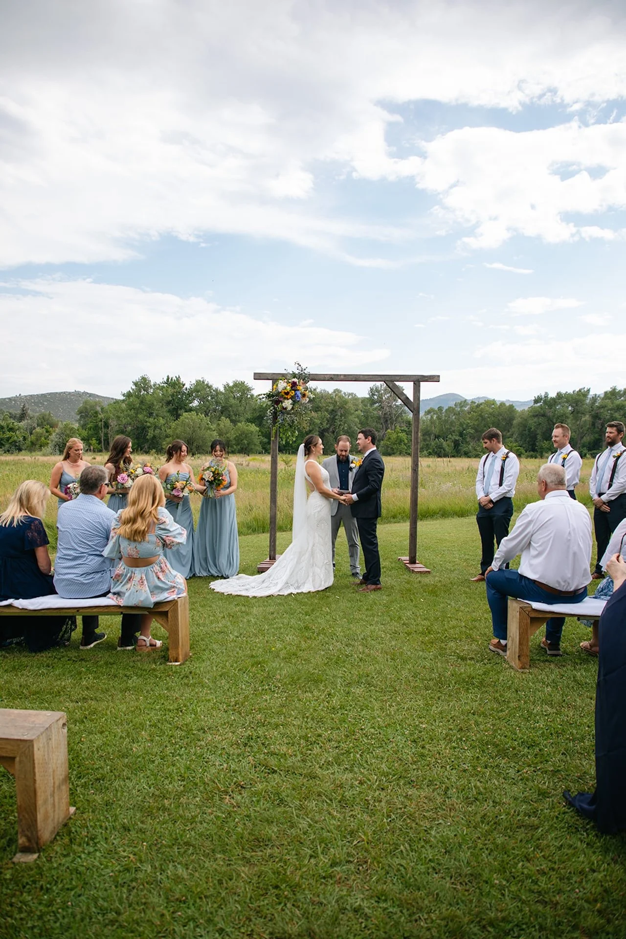 Wide view of an intimate outdoor wedding ceremony with wooden benches, bridesmaids and groomsmen standing on each side, and guests seated in a field with mountain scenery.