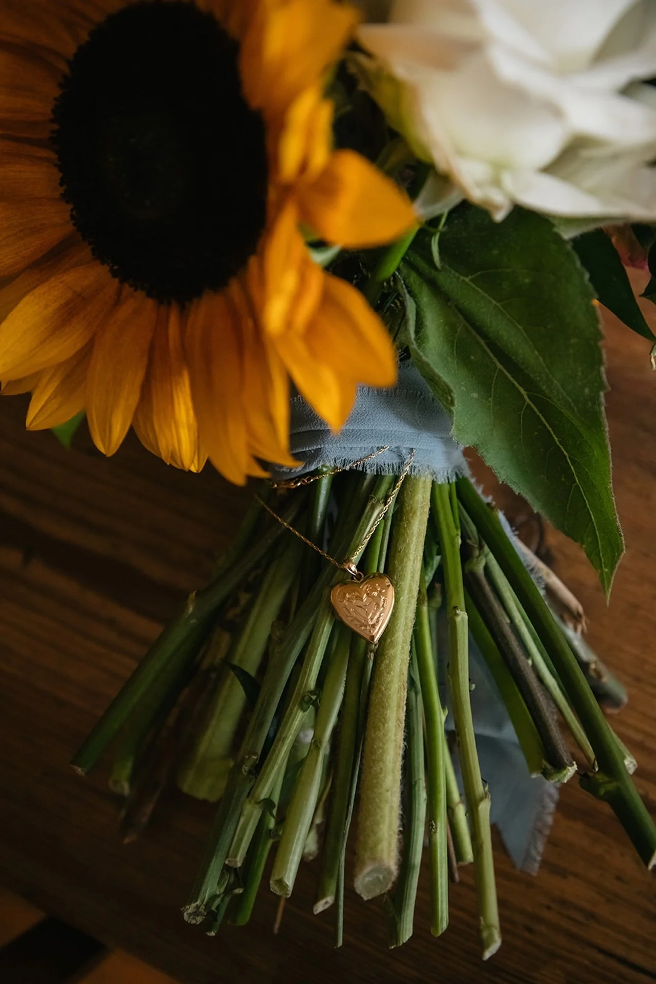 Close-up of a bridal bouquet with sunflowers, greenery, and a heart-shaped gold locket tied with blue ribbon around the stems.