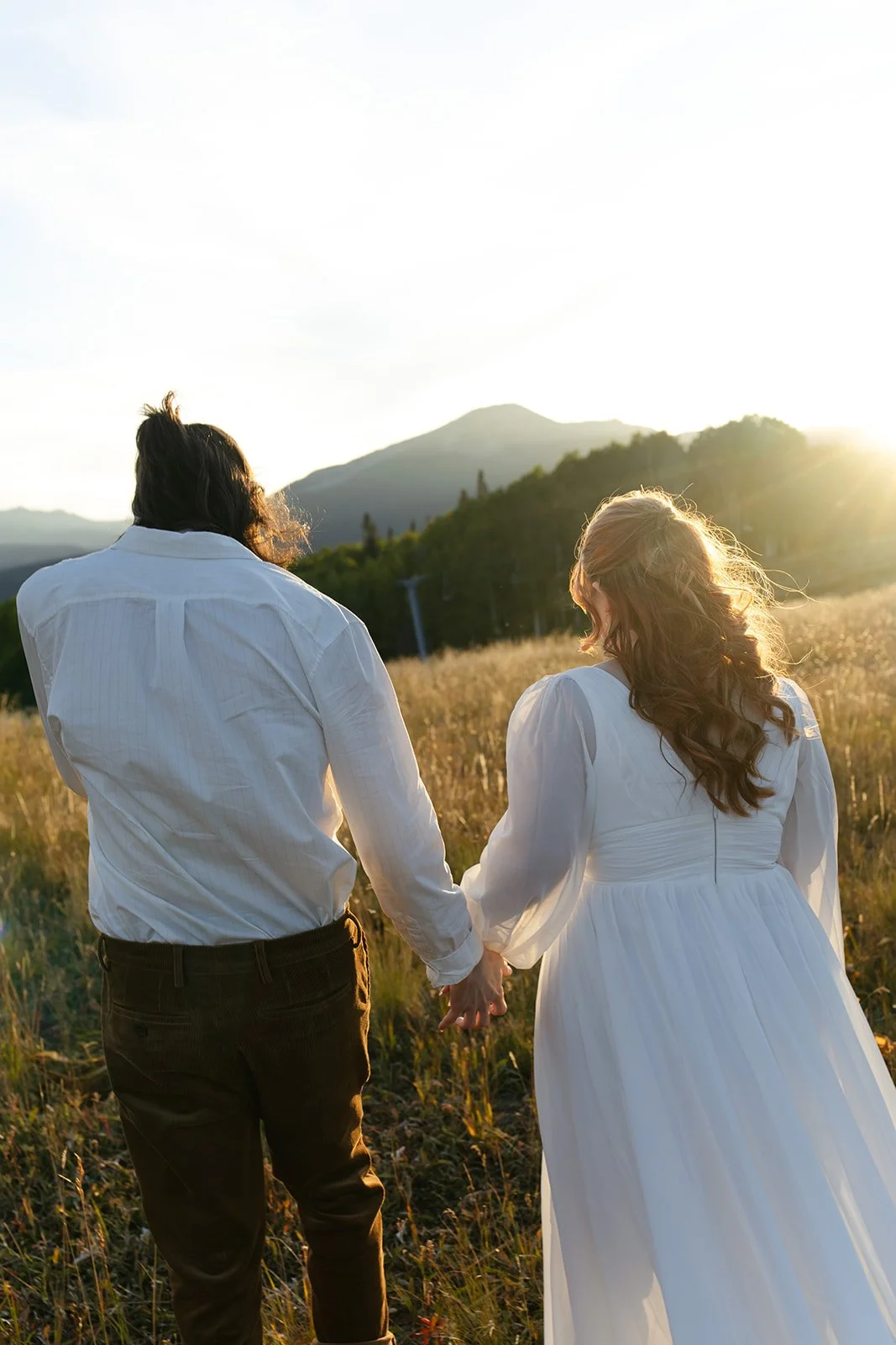 Couple walking hand in hand through a sunlit mountain meadow during their Crested Butte elopement, with rolling hills and soft golden light behind them.
