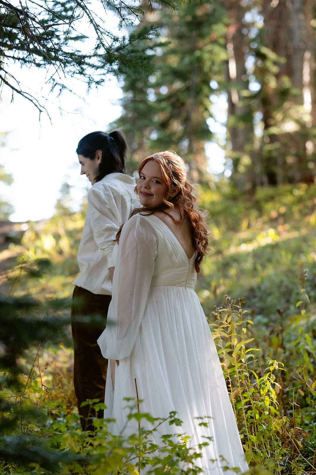 Couple walking hand in hand through a forest trail and the bride looking back at the camera during their Crested Butte elopement.