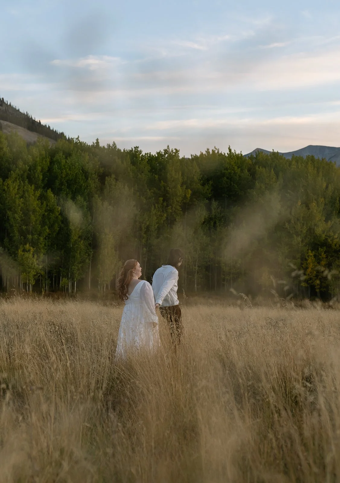 Couple holding hands and walking through tall golden grass during their Crested Butte elopement at sunset with mountain views in the distance.