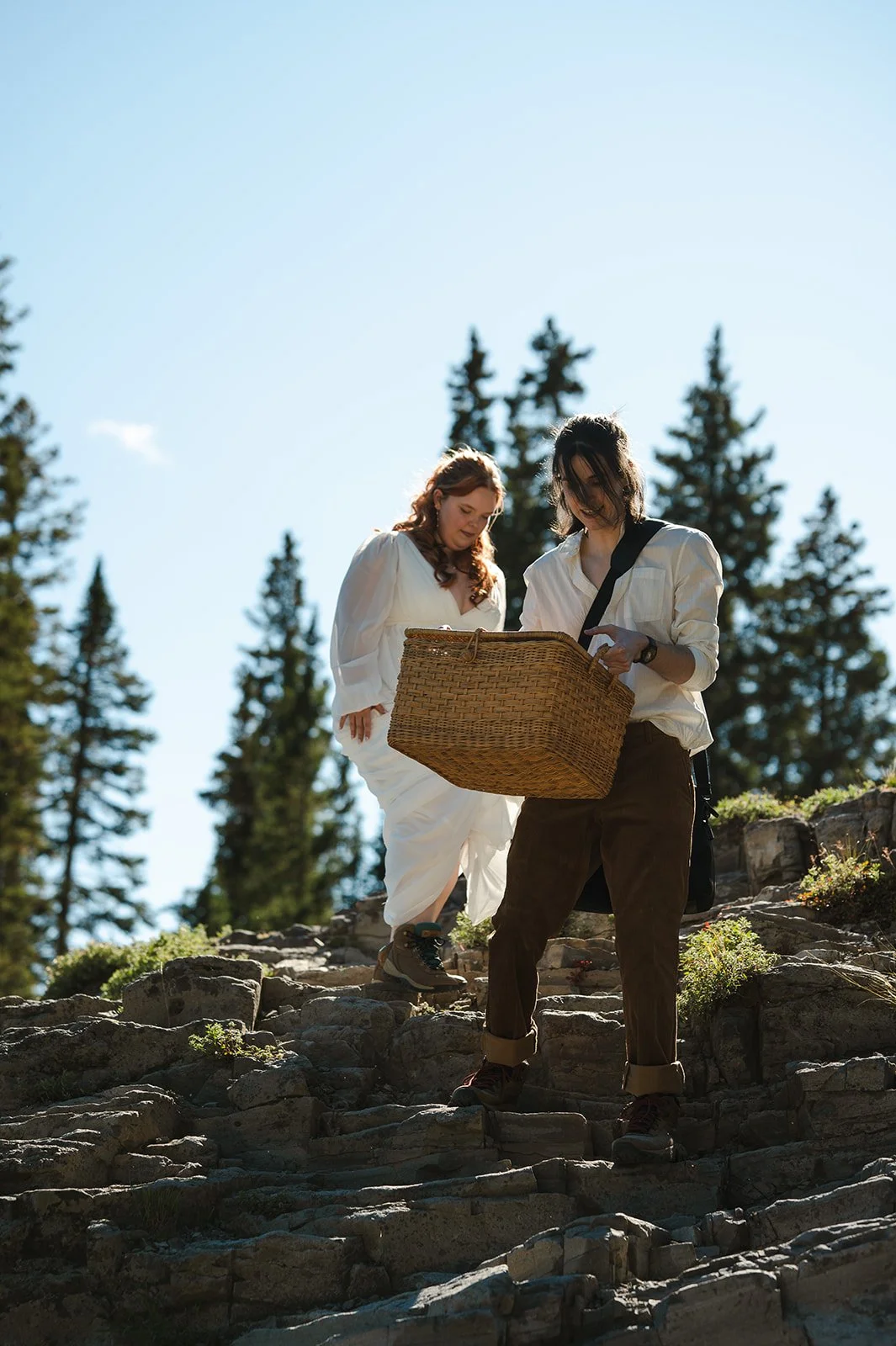 Couple walking along a rocky mountain path during their Crested Butte elopement with evergreen trees and blue sky behind them.