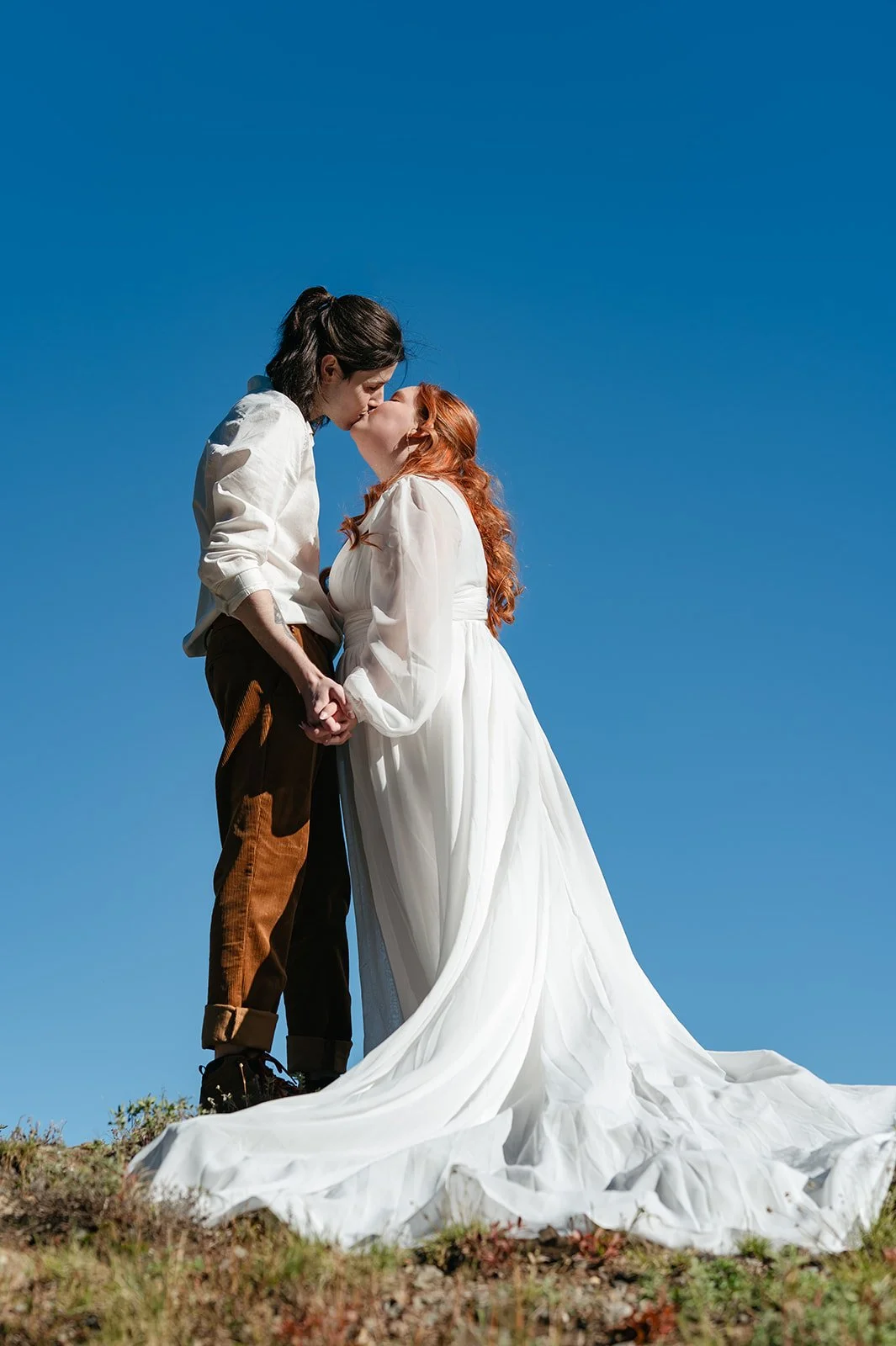 Bride and groom standing on a rocky overlook and sharing a kiss during their Crested Butte elopement, her dress flowing dramatically in the wind against a clear blue Colorado sky.