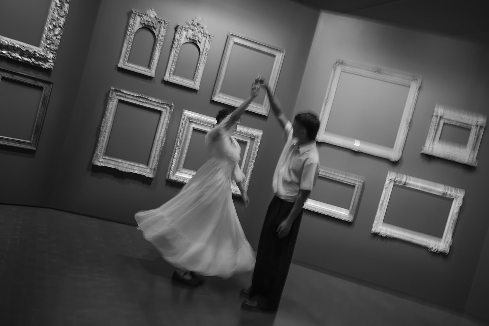 A romantic black and white scene of a couple dancing together in front of a wall filled with empty frames, emphasizing motion and connection.