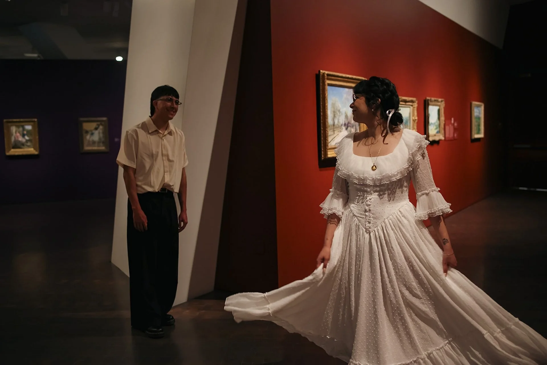 A romantic moment of a couple facing each other inside a museum gallery, separated by a wall opening, with framed artwork and warm red walls creating cinematic city engagement photos.