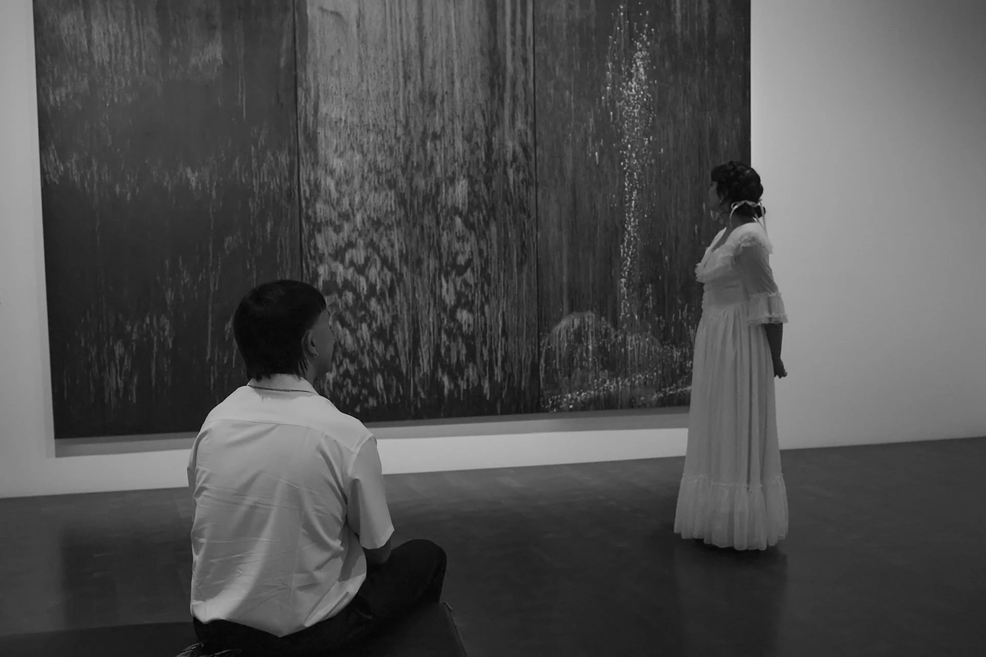 A black and white image of the man seated on the floor while his fiance stands across the gallery, both focused on a large abstract artwork.