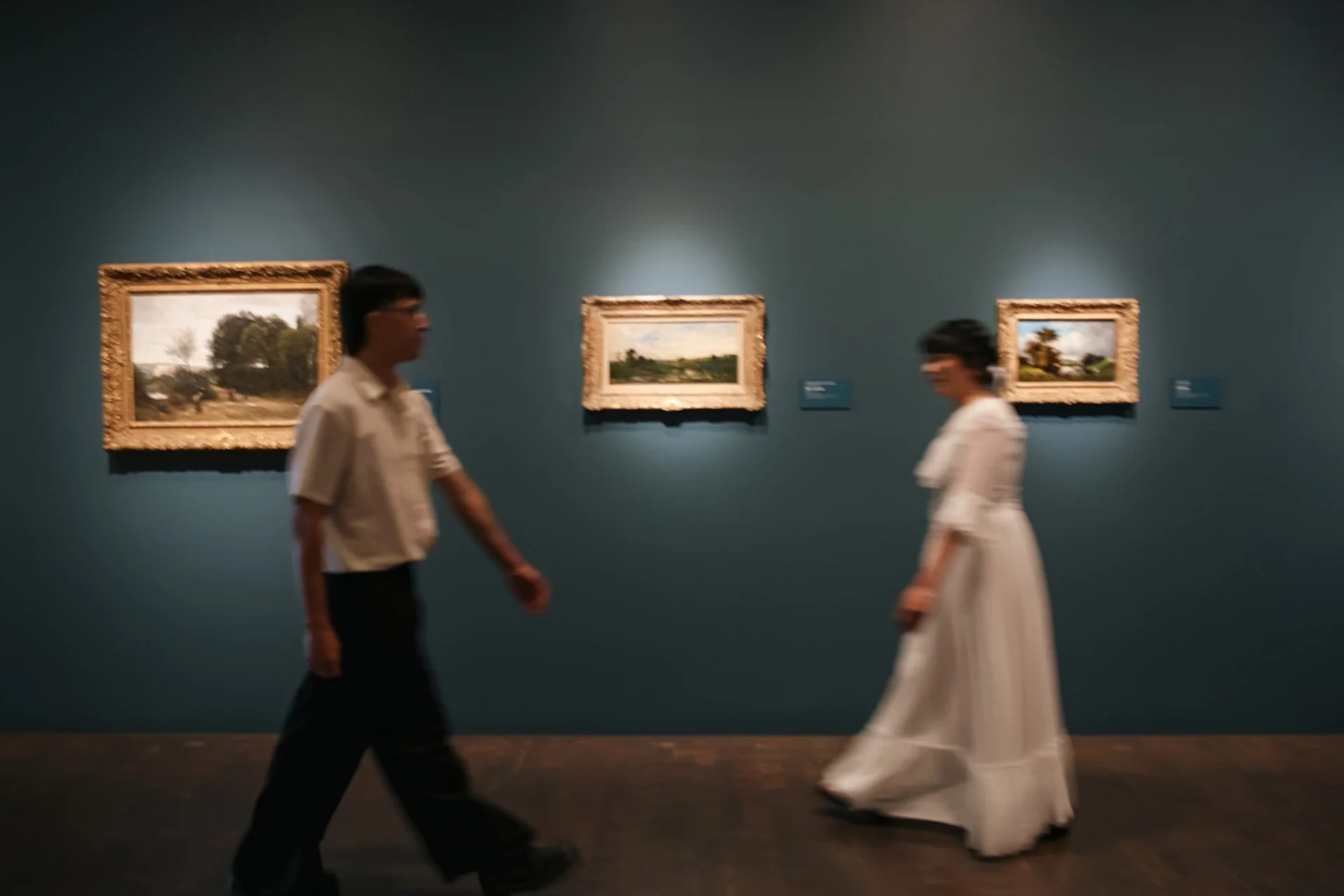 A wide museum gallery shot of a couple walking toward each other between framed landscape paintings, emphasizing movement and storytelling.