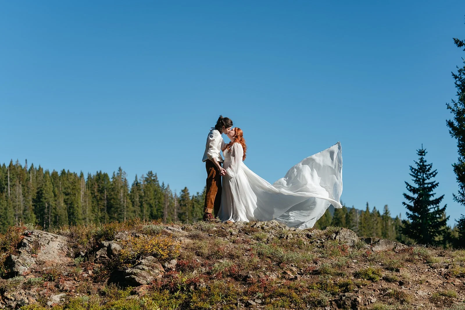 Bride and groom standing on a rocky overlook during their Crested Butte elopement, her dress flowing dramatically in the wind against a clear blue Colorado sky.
