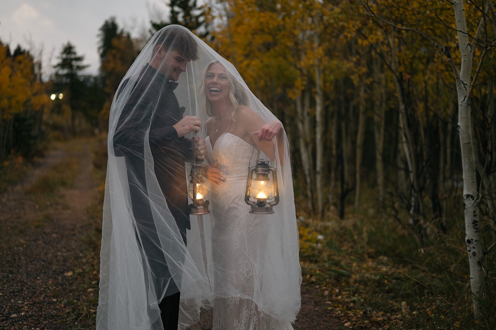 a bride and groom laughing together underneath a veil as they hold lanterns in CO