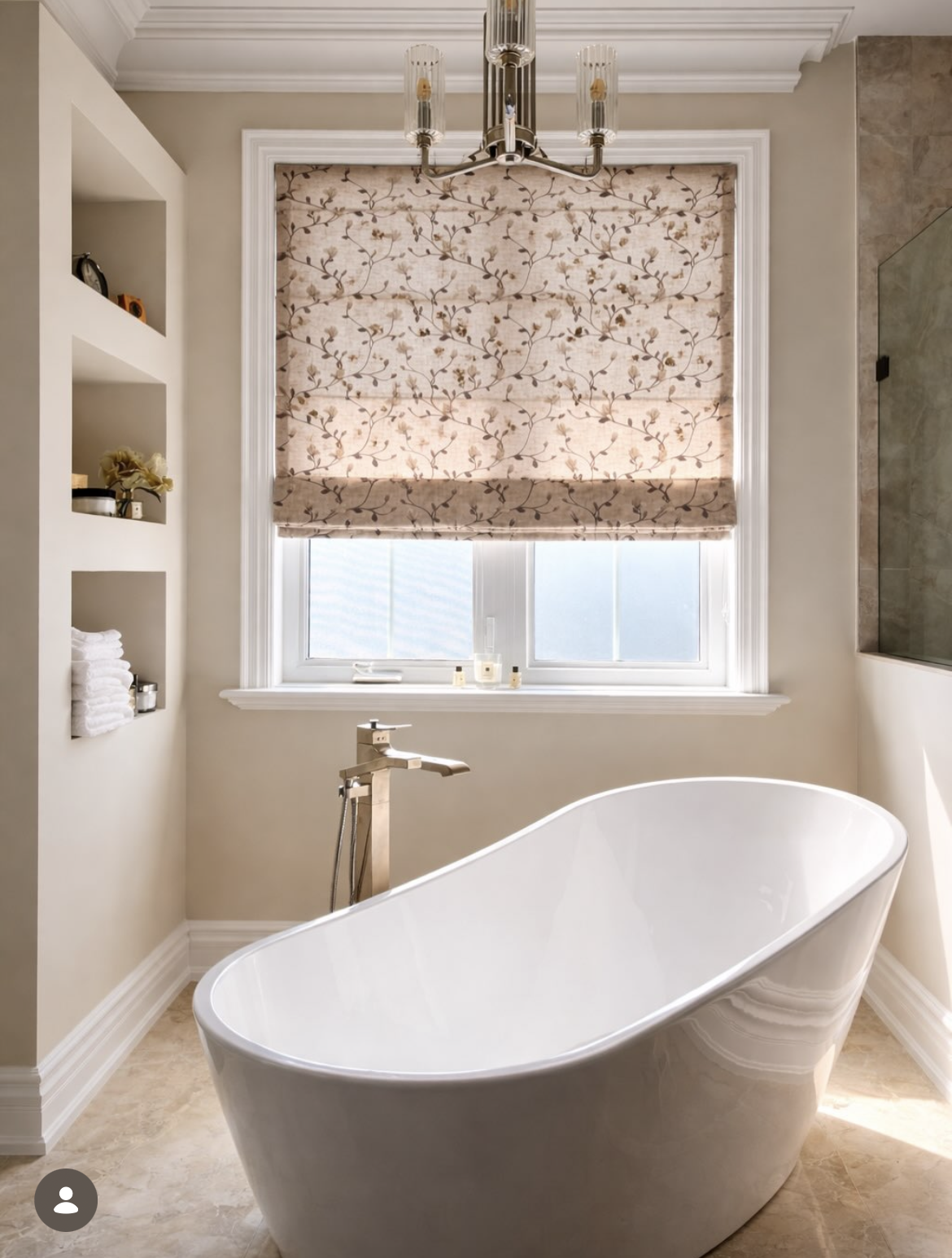 Modern bathroom with a freestanding bathtub, window with patterned Roman shade, built-in shelves with towels and decorations, chandelier, and marble flooring.
