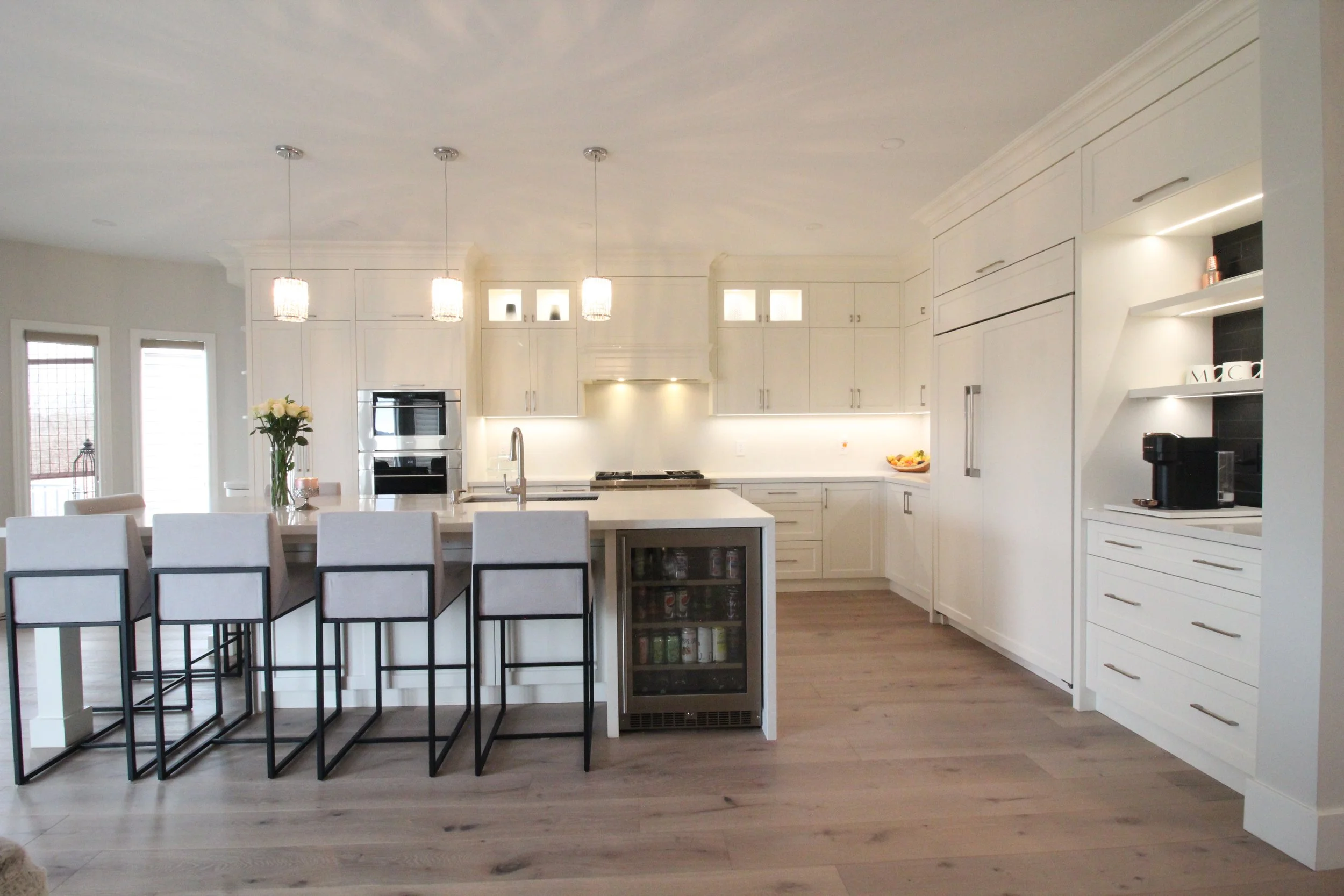 Modern kitchen with white cabinets, a center island, hanging pendant lights, and stainless steel appliances. There are large windows in the background, and a vase of white flowers on the island.