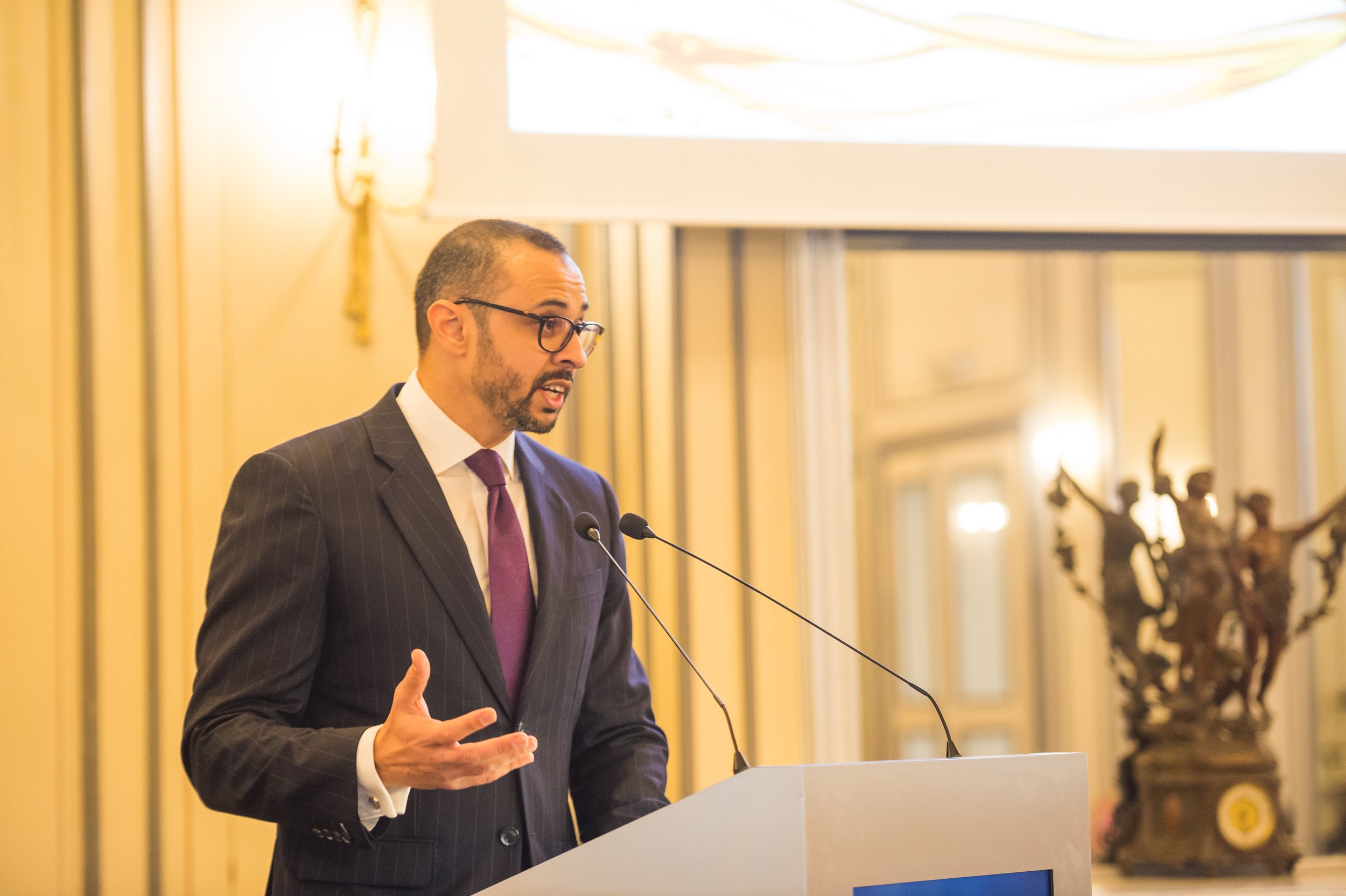 Man in a business suit with glasses speaking at a podium in a formal room with classical decor.