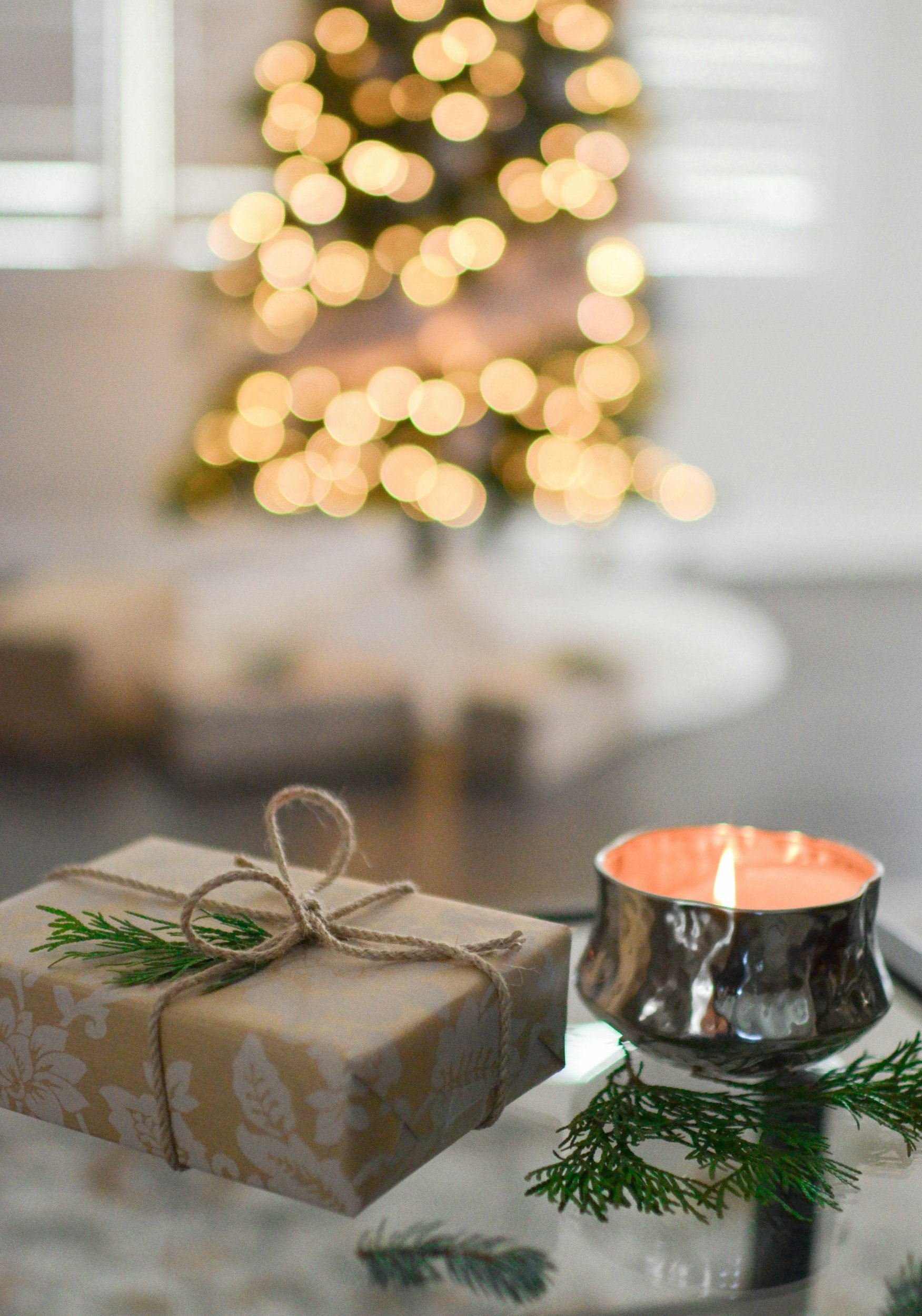 A wrapped Christmas gift with a sprig of evergreen and a candle in a decorative holder on a table, with a blurred Christmas tree with lights in the background.