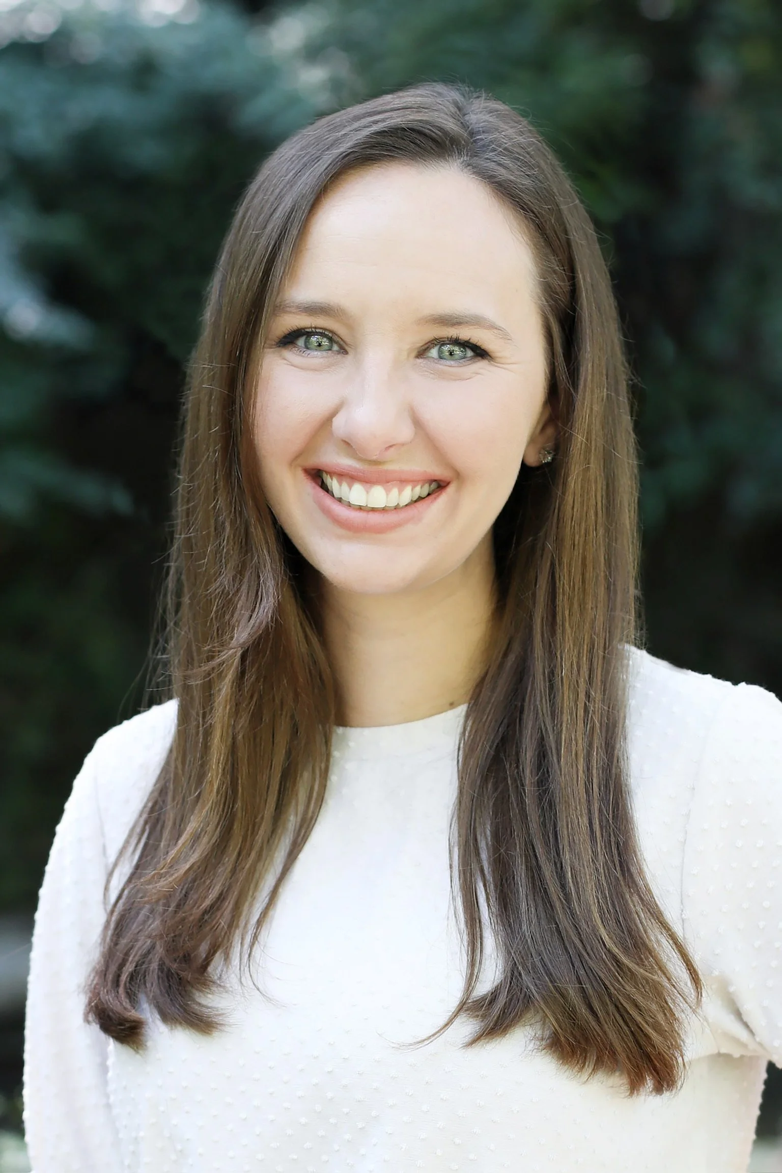 A young woman with long brown hair and green eyes, smiling outdoors in a white sweater with a textured pattern, with green foliage in the background.