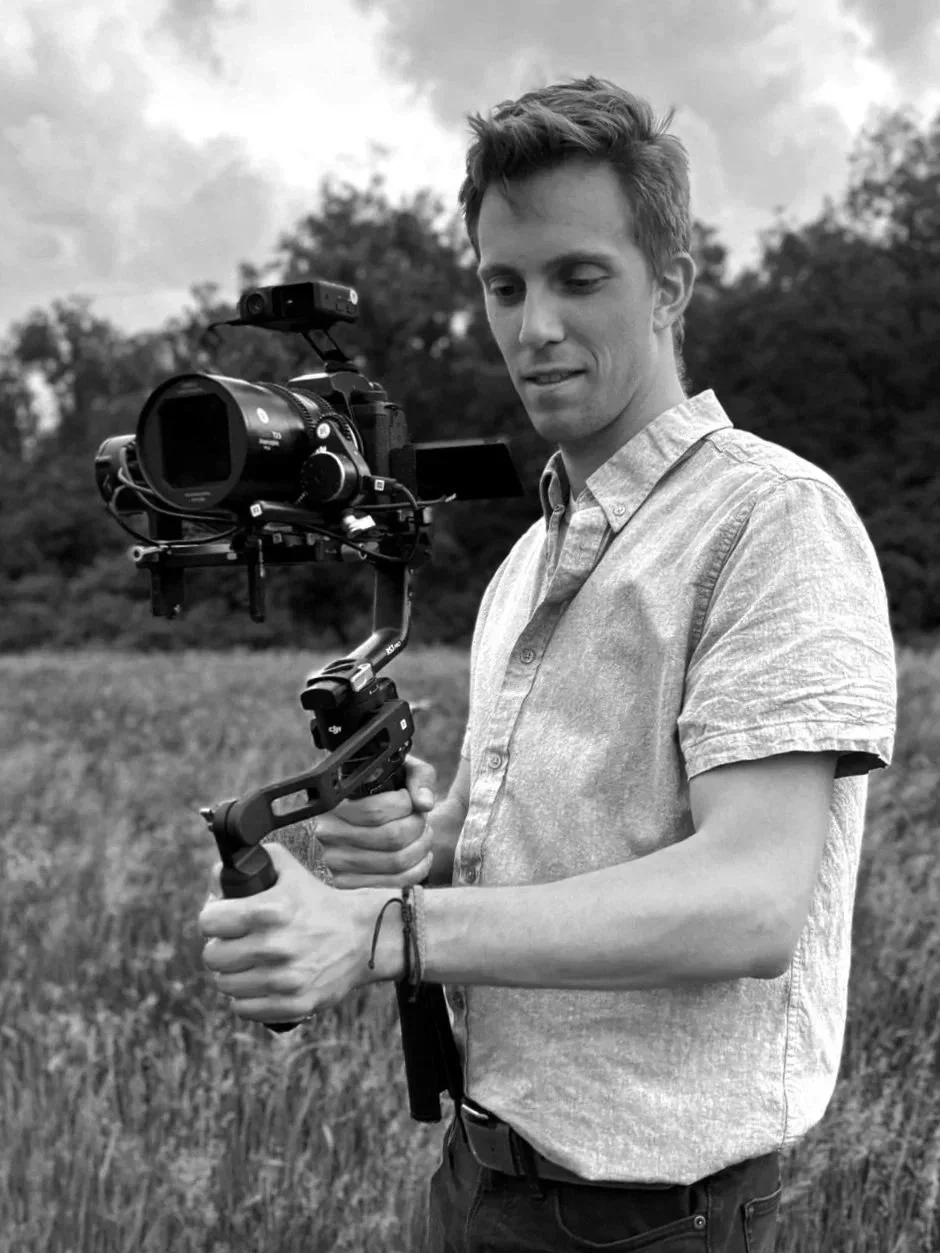 A young man outdoors holding a professional camera stabilizer rig and camera, with trees and sky in the background, in black and white.