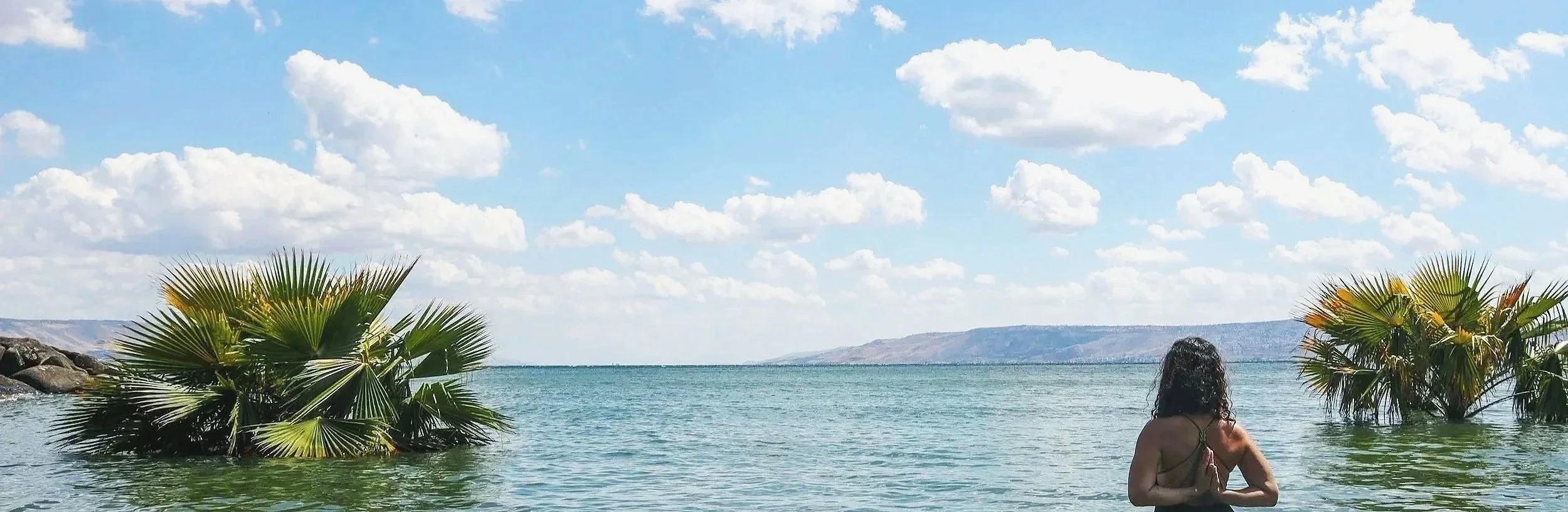 A woman standing in a lake or pond surrounded by palm trees, with a landscape of hills and a blue sky with clouds in the background.