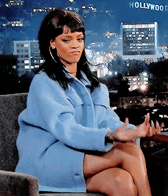 Rihanna on a late-night talk show makes money sign gestures with both hands while seated in a blue coat, reacting confidently against a Hollywood city skyline backdrop.