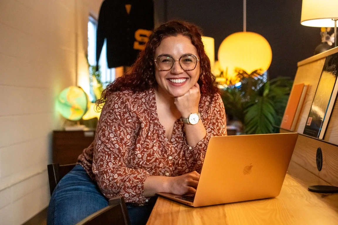 Raven smiling working on a laptop in a cozy Switchyards Club with warm lighting, plants, and modern decor, representing small business productivity.