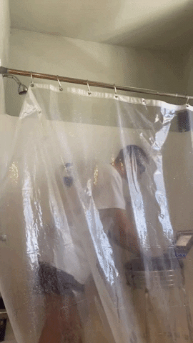 Person standing fully clothed in a running shower behind a clear plastic curtain, clothes soaked, typing on a laptop on a barstool while brushing their teeth in a small bathroom.