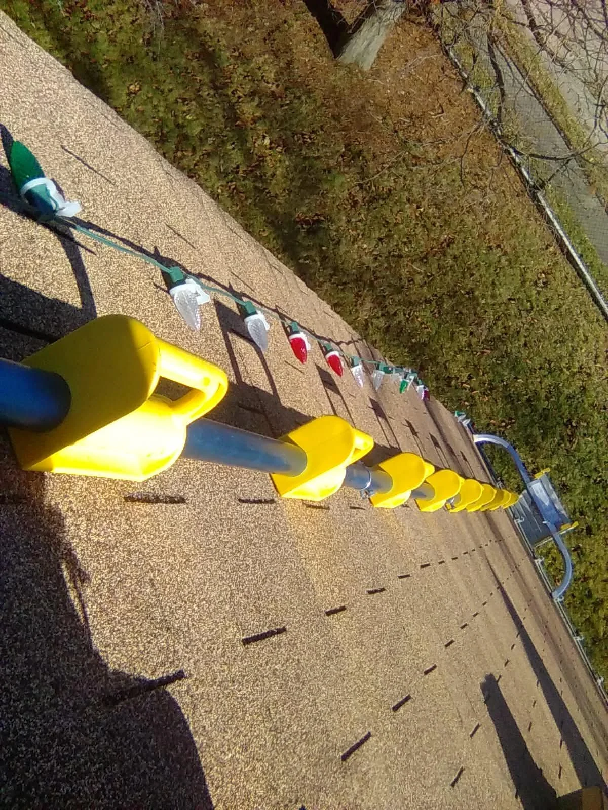 A row of yellow creative barriers lining the edge of a road, with a string of holiday lights hanging on a rope attached to the barriers. Trees with brown and green leaves are visible in the background, indicating it is autumn.