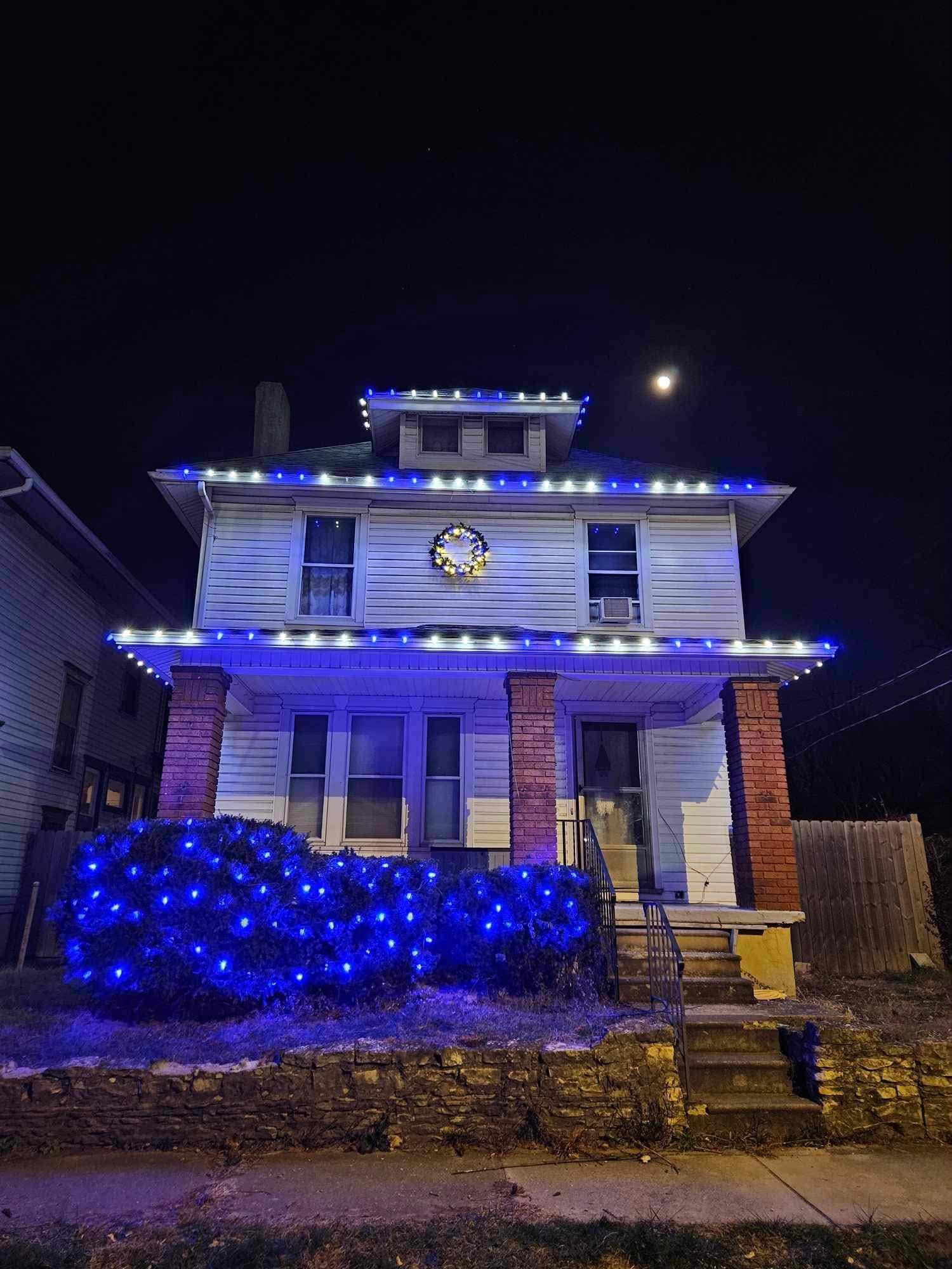 A two-story house decorated with blue and white Christmas lights, with a wreath on the front wall, and a bush illuminated with blue lights in the front yard at night. The moon is visible in the dark sky.