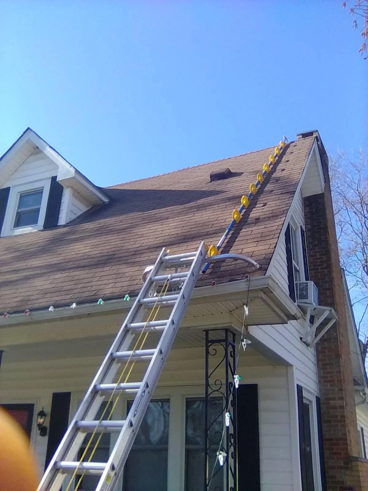 The image shows a house with a ladder leaning against the roof with yellow clips attached to a blue pole, likely for installing Christmas lights. There are also string lights hanging from the house's eaves.