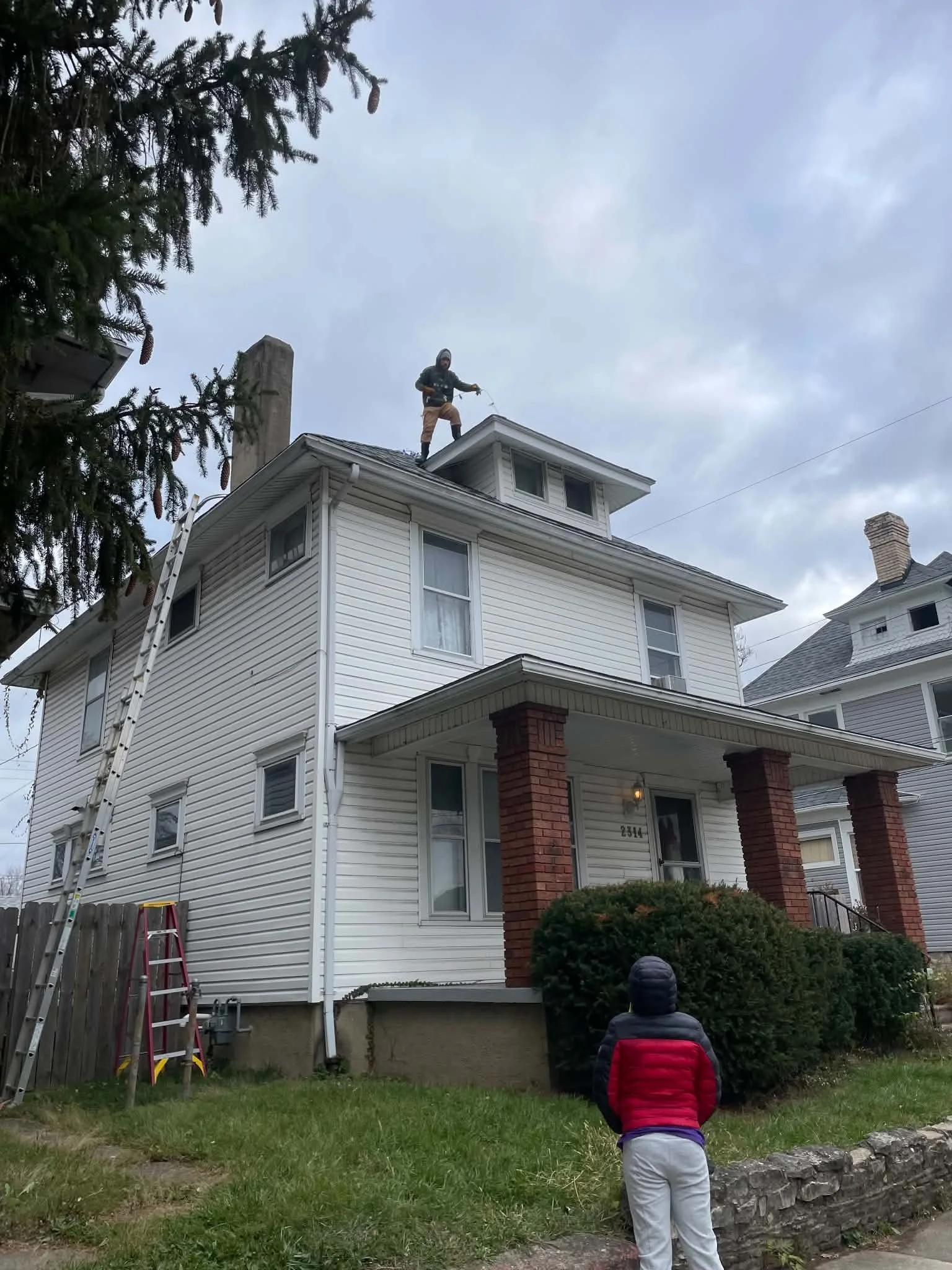 A person standing on the roof of a white house in a residential neighborhood, holding a cable or rope. A child is standing on the lawn looking up at the house. The house has multiple stories, a chimney, and a ladder leaning against it. There are trees and other houses nearby.
