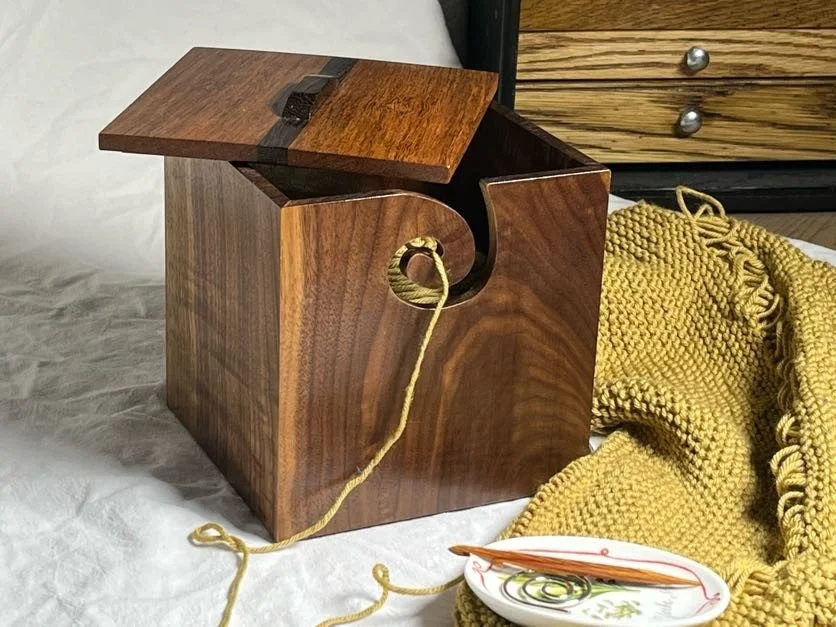 A yarn box holding a mustard colored yarn that is attached to a knitting project. 