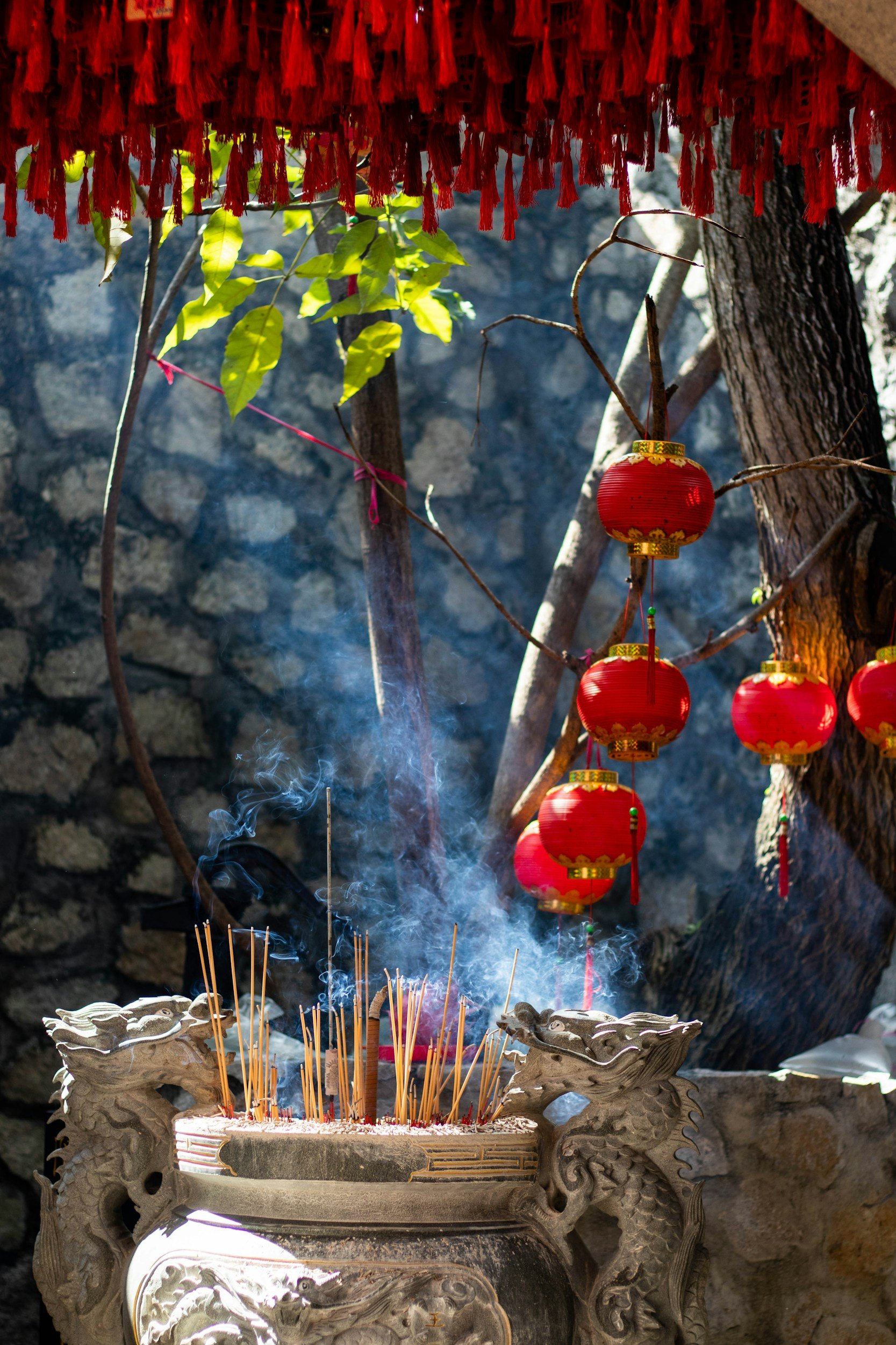 Red Chinese lanterns hanging above an incense burner with burning incense sticks, smoke rising, outdoors near trees and rocks.