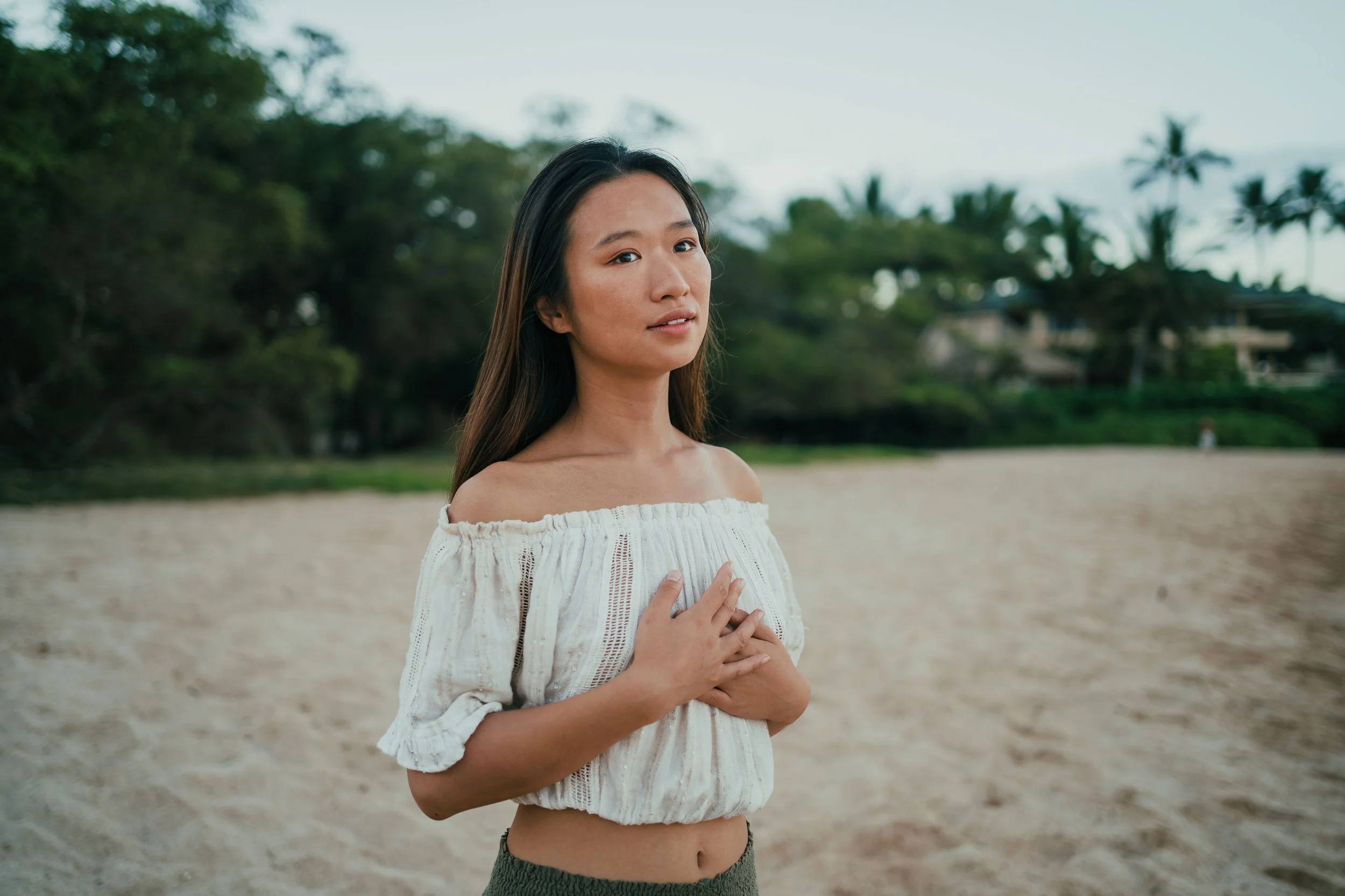 A woman standing on a beach with trees and houses in the background, wearing an off-shoulder white top and holding her hands over her chest.